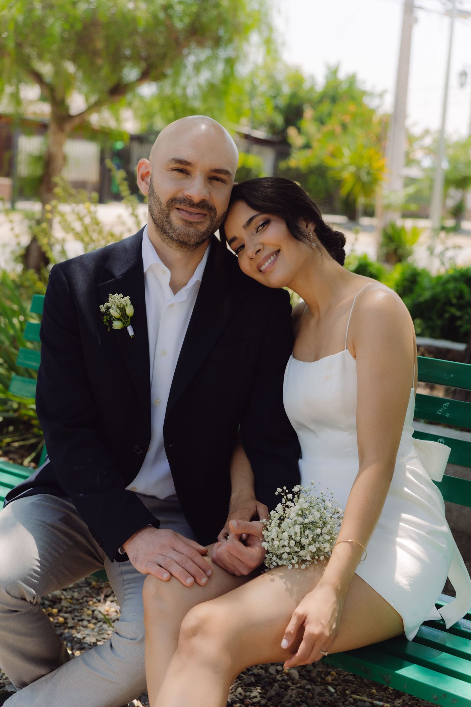 Smiling newlyweds sit closely together on a green park bench holding delicate white flowers.