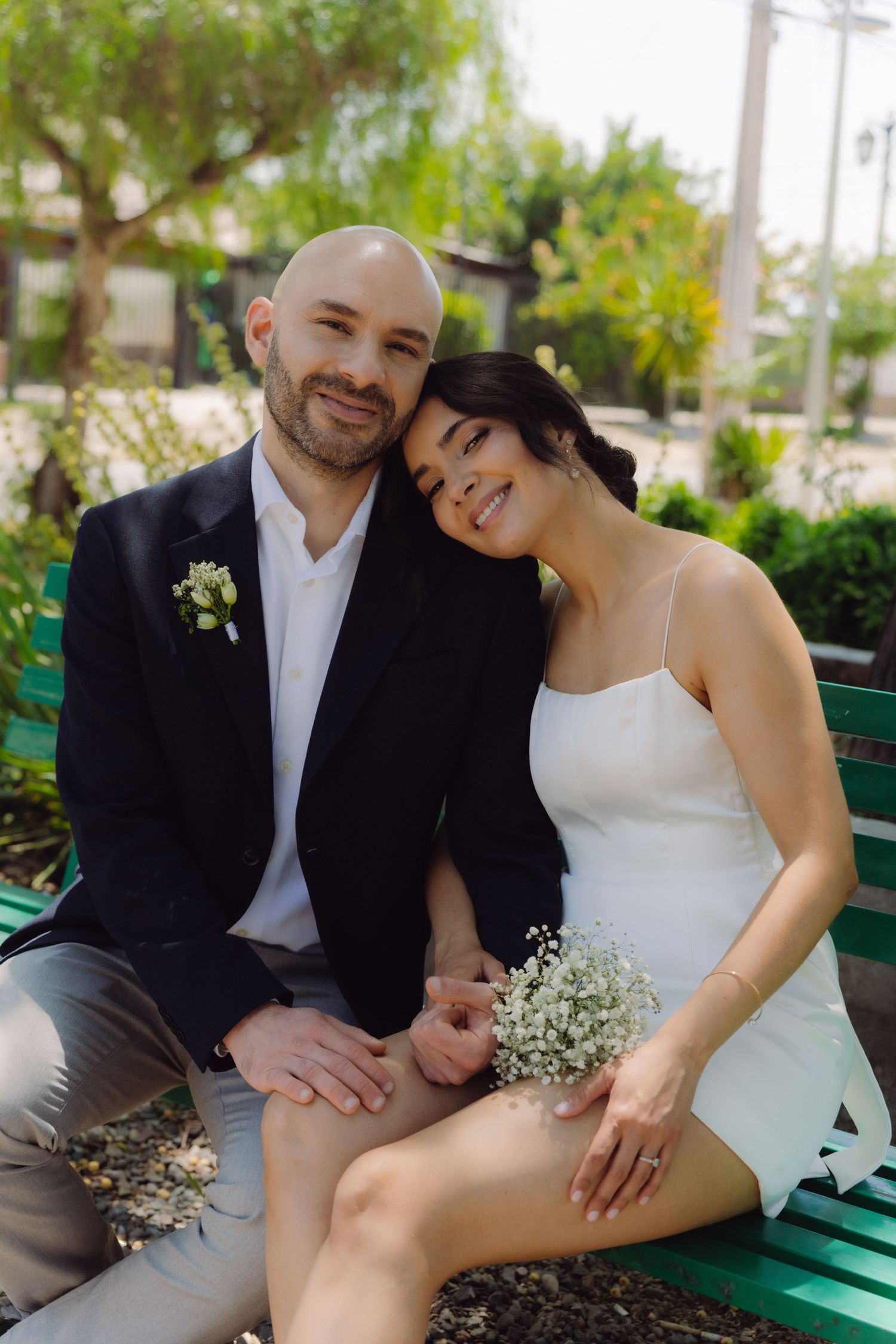 Happy bride leans on groom's shoulder while sitting on a bench holding baby's breath bouquet.