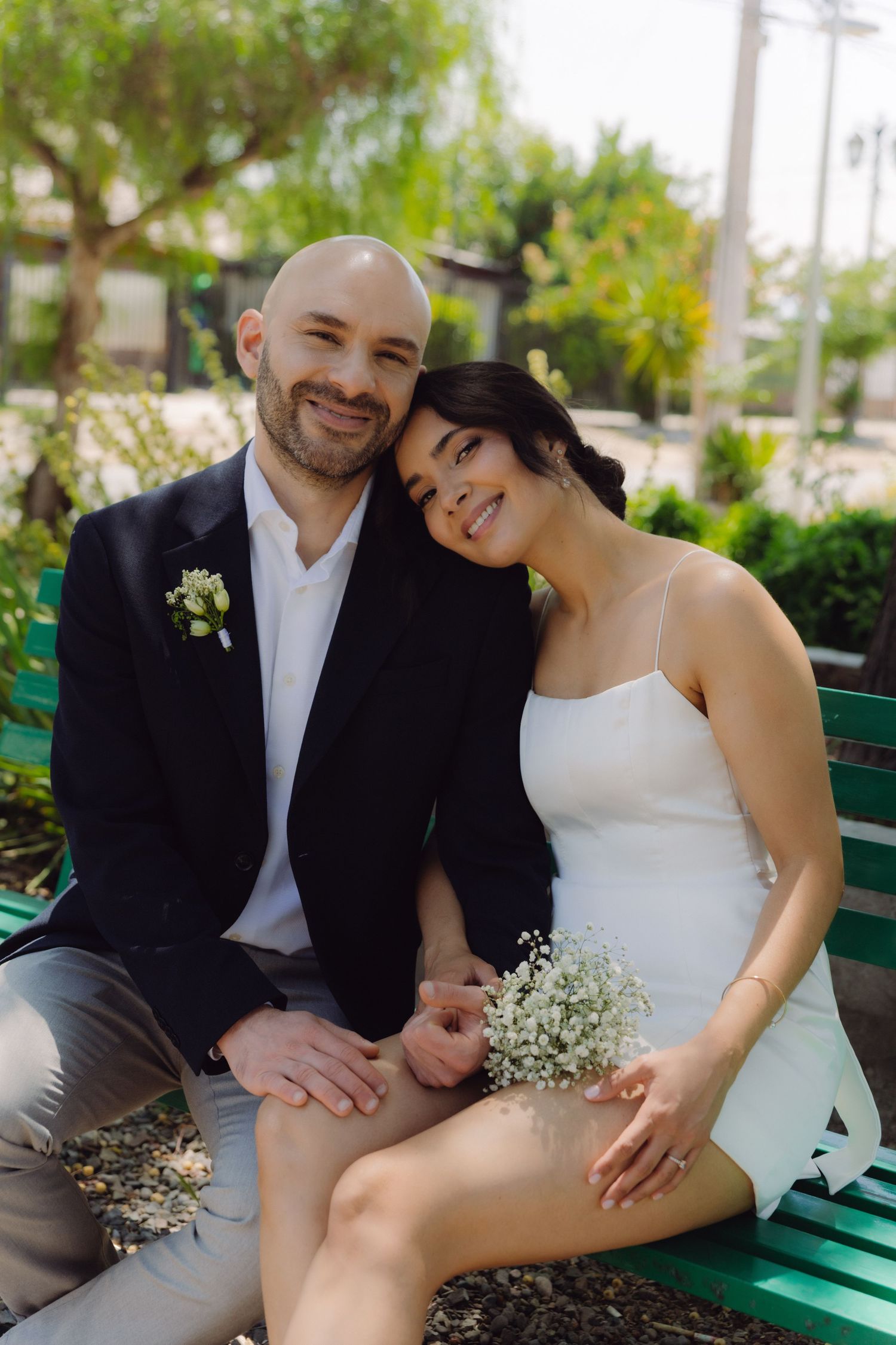 Smiling bride and groom sitting on a green bench outdoors, bride holding baby's breath bouquet in wedding attire.