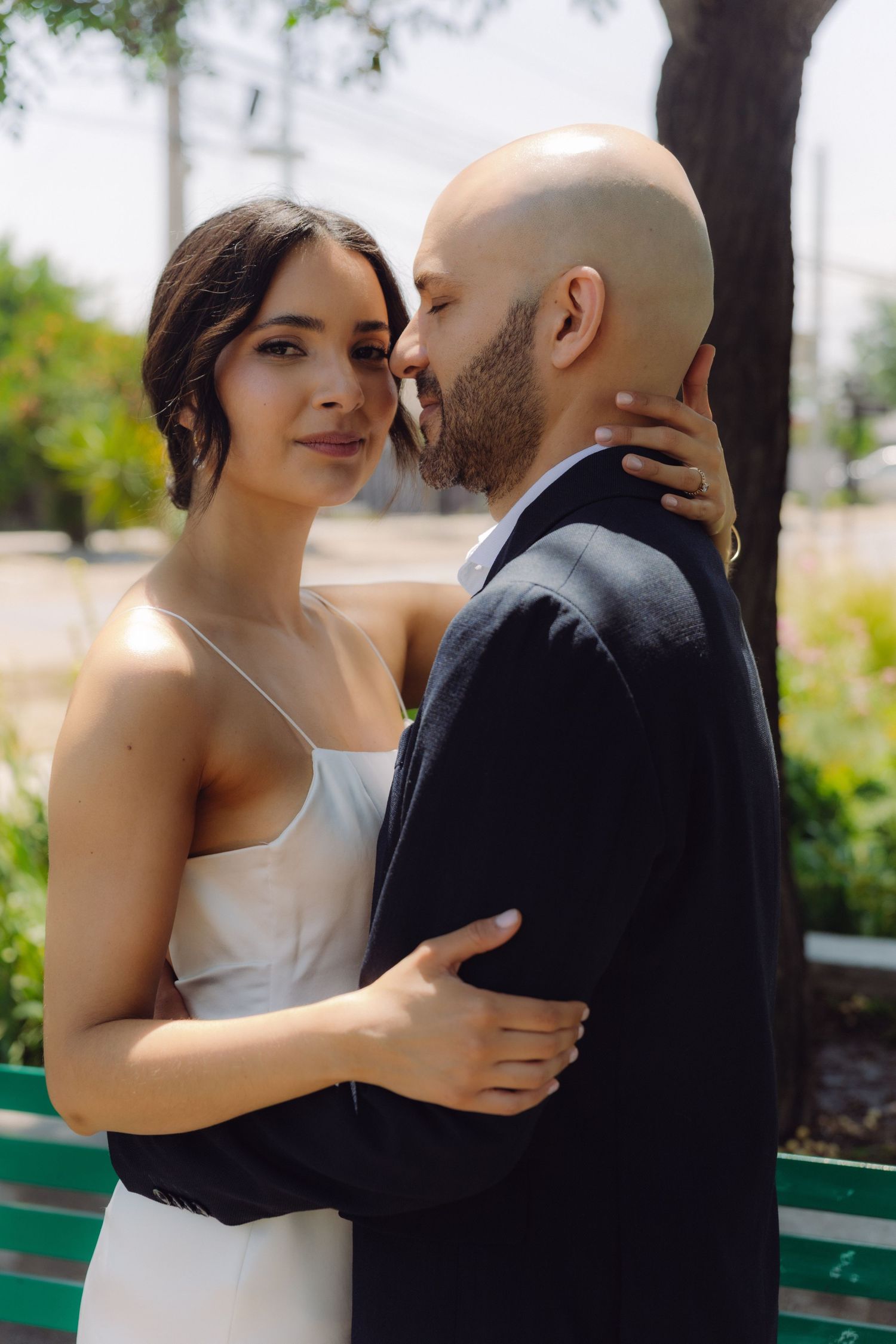 Bride and groom embracing outdoors, bride in white mini dress looking at camera while groom kisses her cheek.