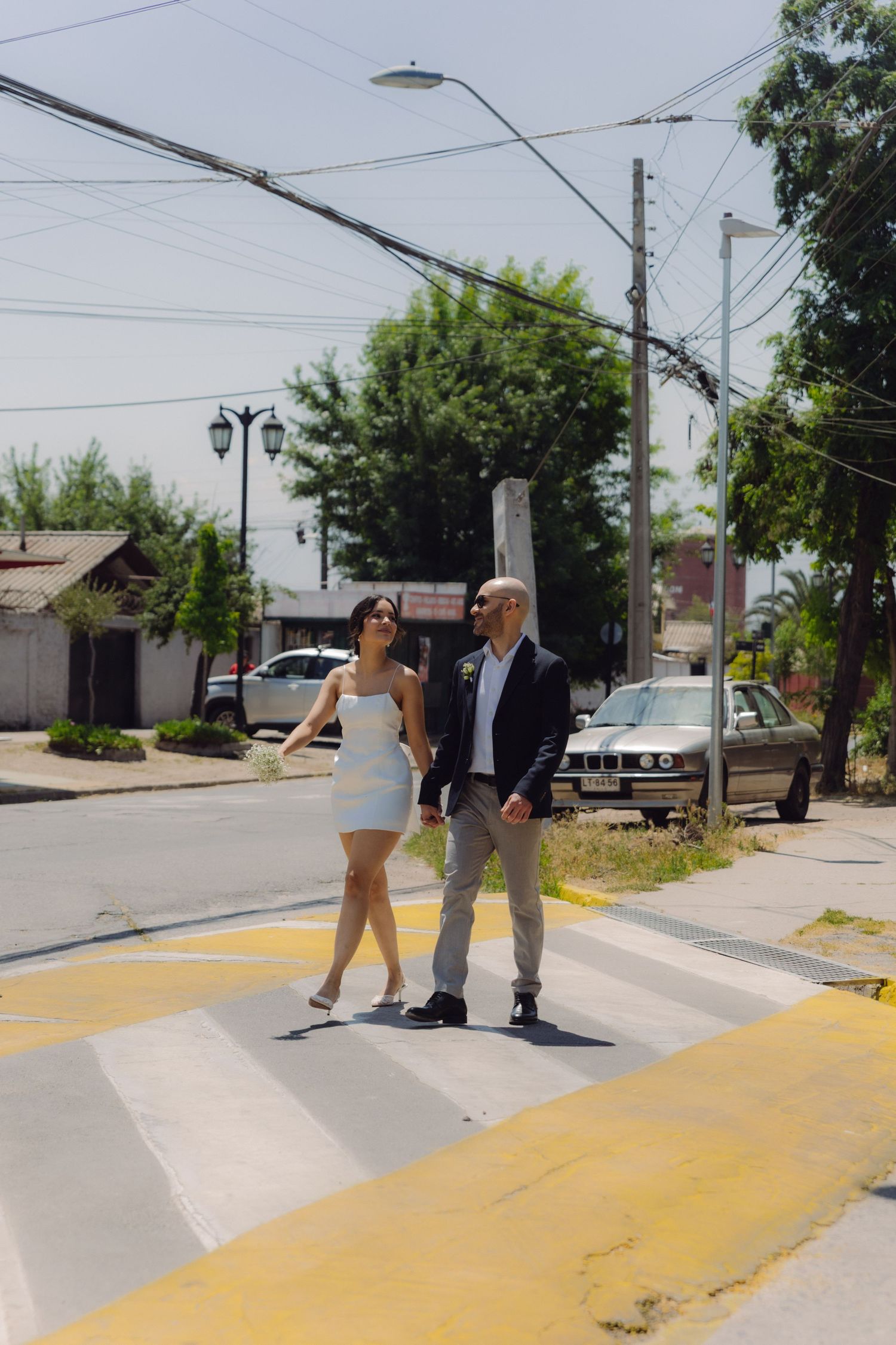Bride and groom holding hands while crossing a zebra crosswalk on a sunny street in wedding attire.