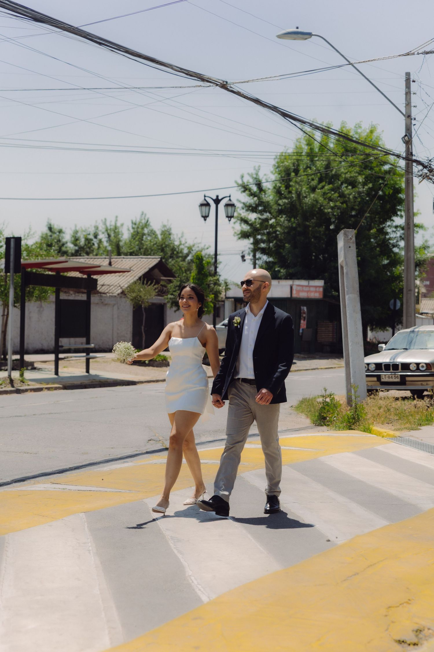 Newlywed couple walking hand in hand across a yellow and white pedestrian crosswalk on a bright sunny day.