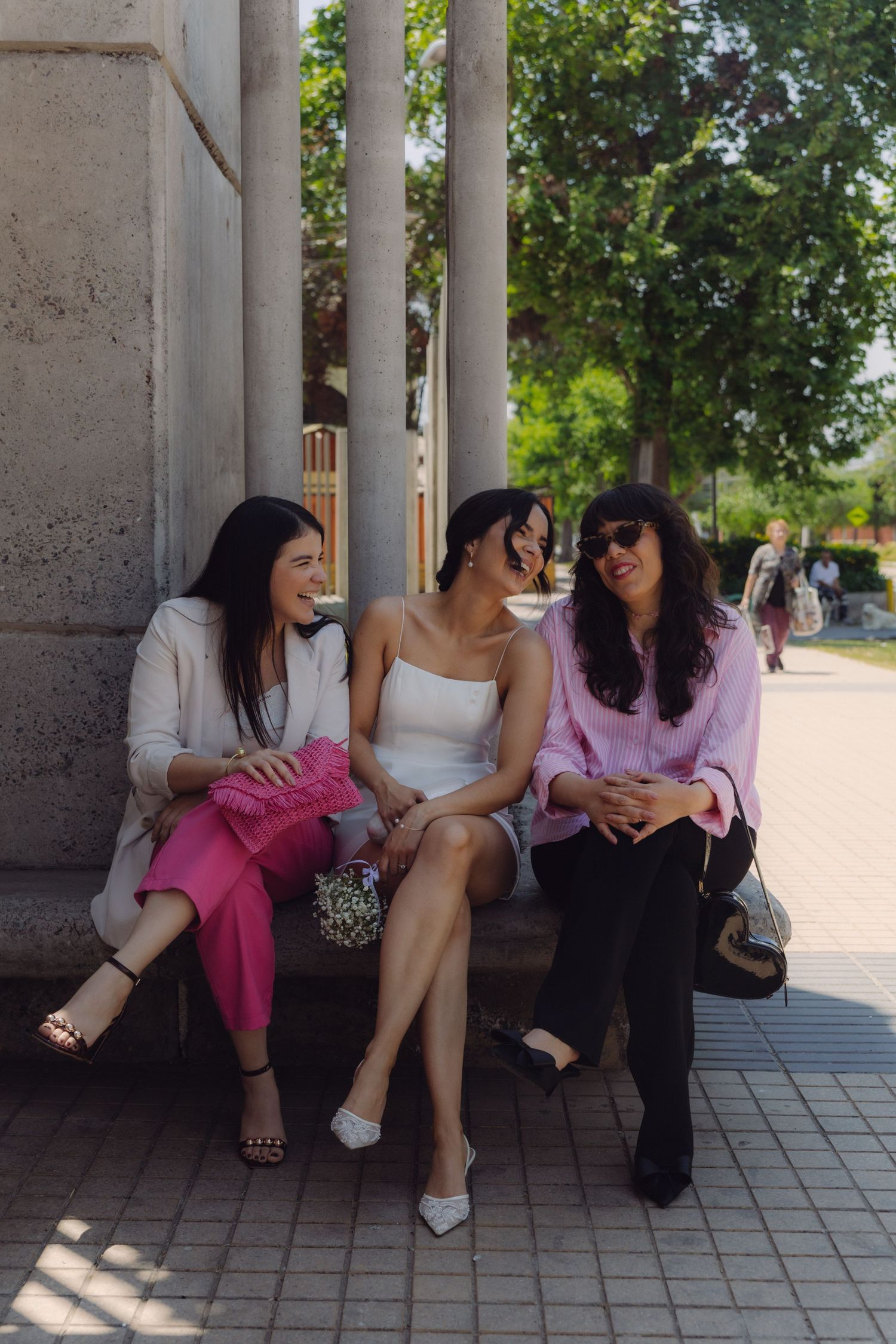 Bride in white mini dress sitting and laughing with two female guests in pink outfits beside stone columns.