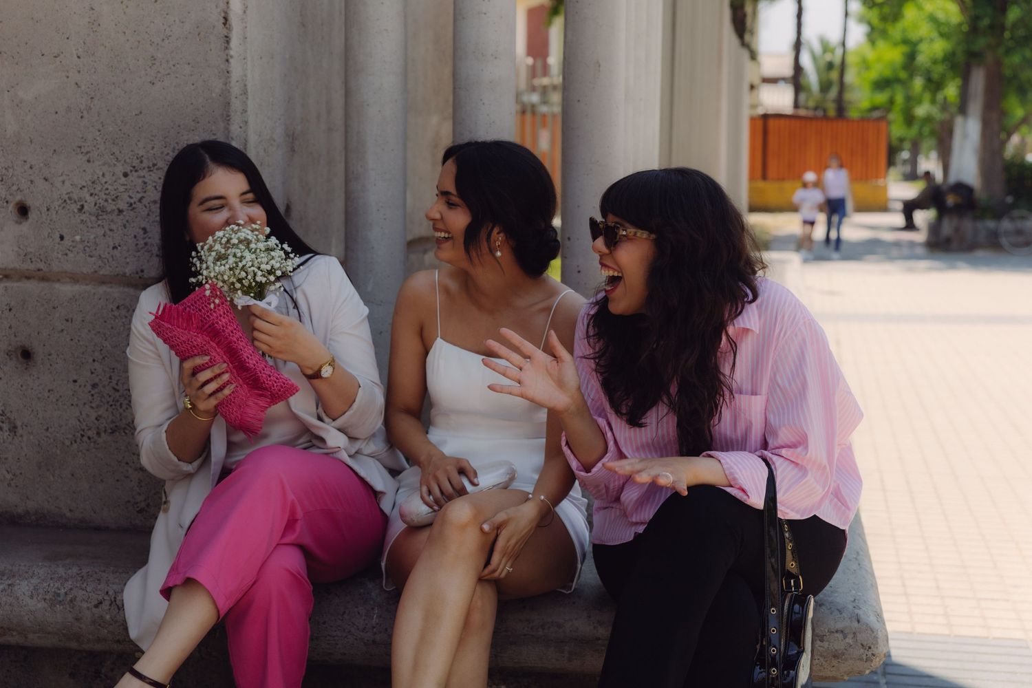 Bride and two friends sitting on steps laughing, one friend holding a pink clutch and baby's breath bouquet.