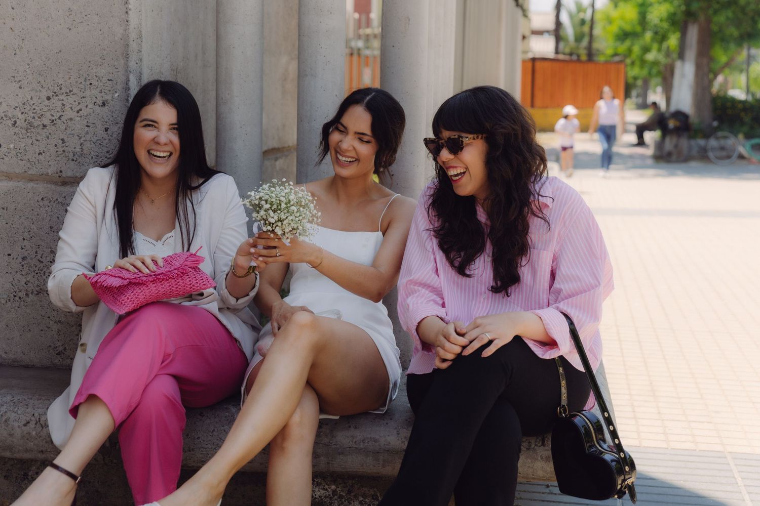 Three women sitting outdoors laughing, bride in white holding floral bouquet surrounded by friends in pink.