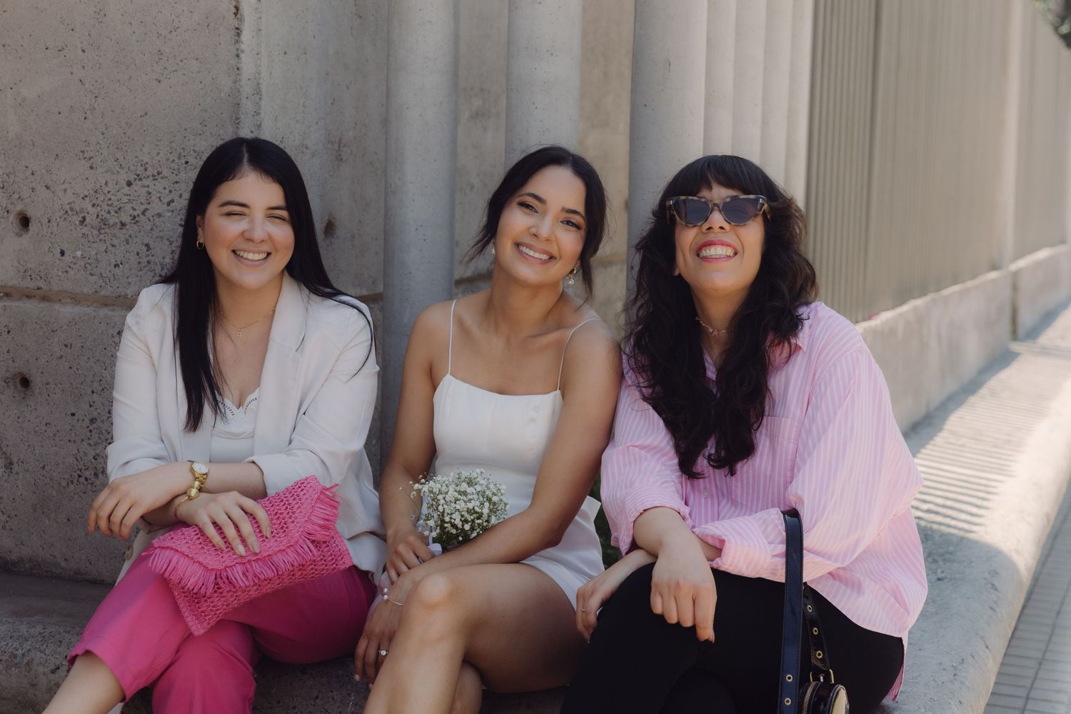 Bride in white dress posing with two smiling female guests beside stone columns on a sunny day.