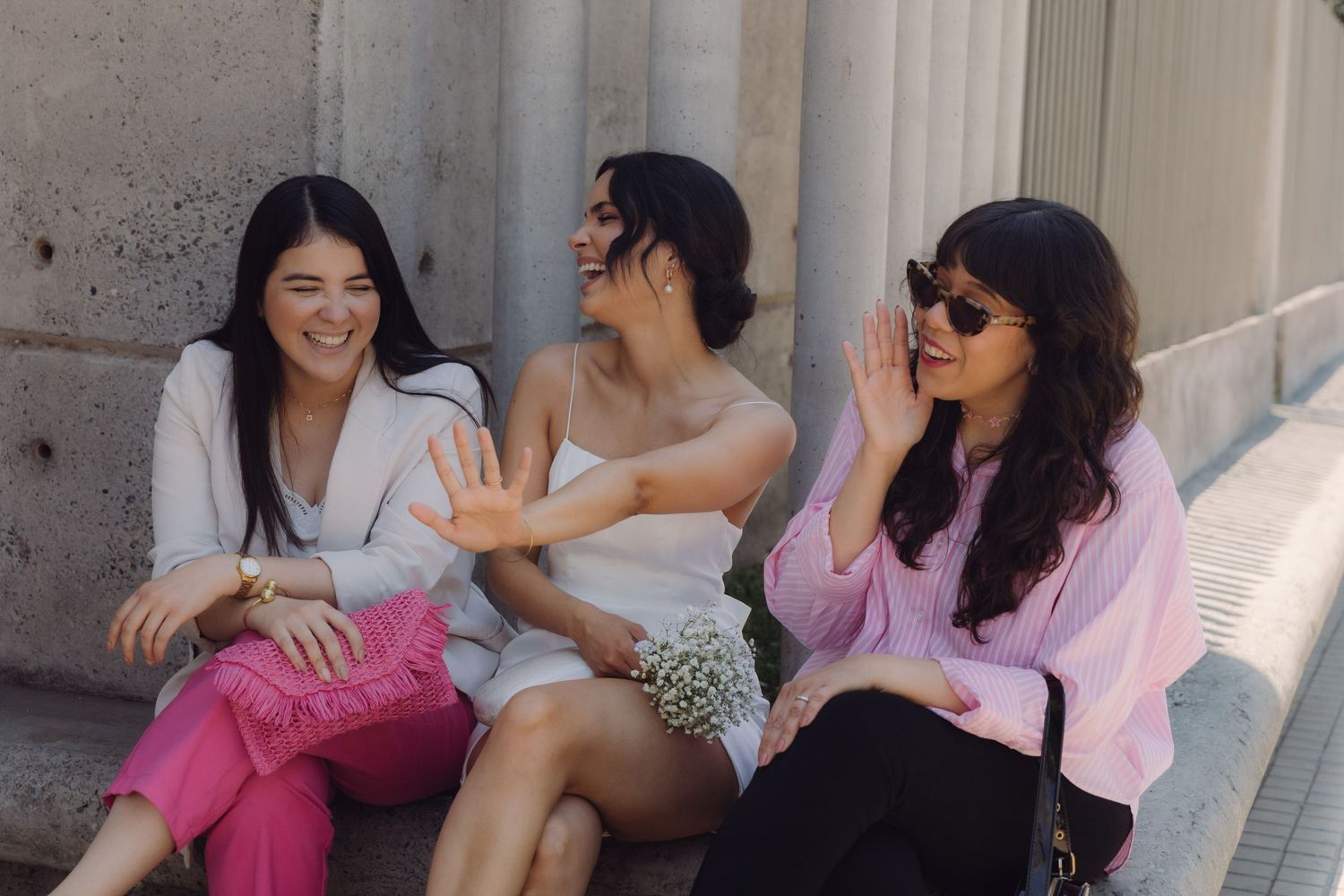 Three women sitting on steps laughing and chatting outdoors, one in white dress holding flowers.