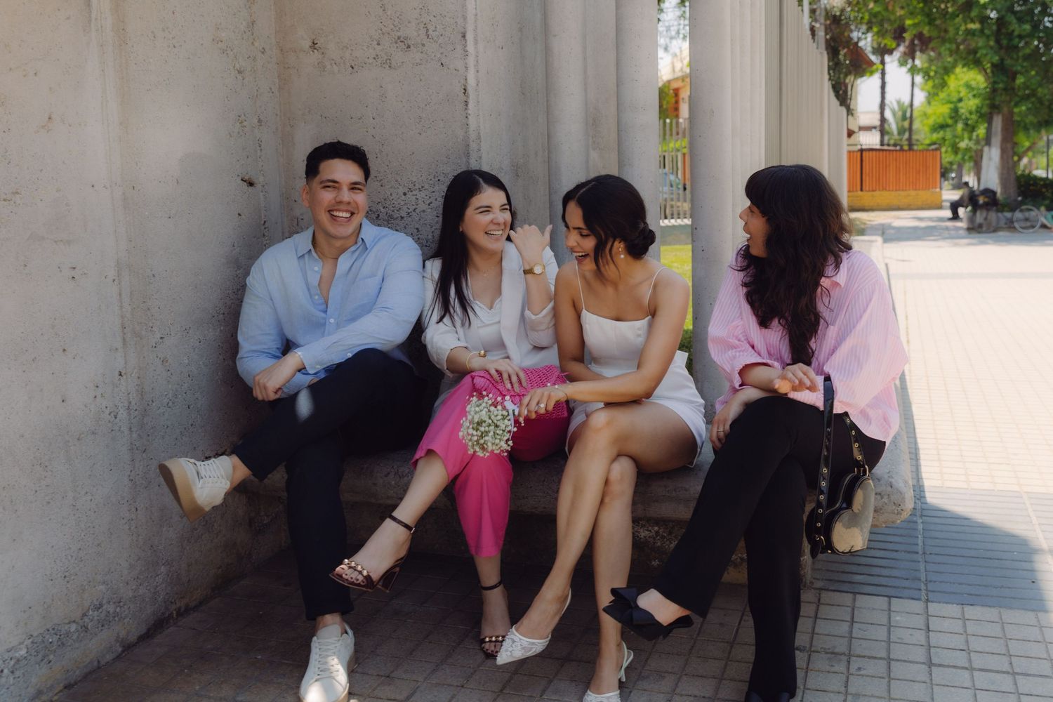 Four young adults sitting and laughing together outdoors against a concrete wall in sunlight.