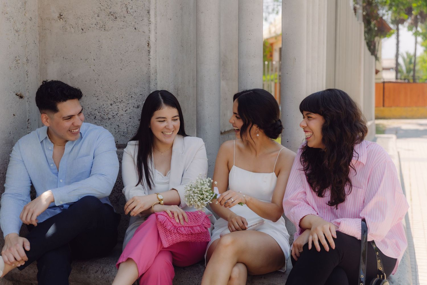 Four friends sitting outdoors on steps, smiling and holding small white flowers in sunlight.