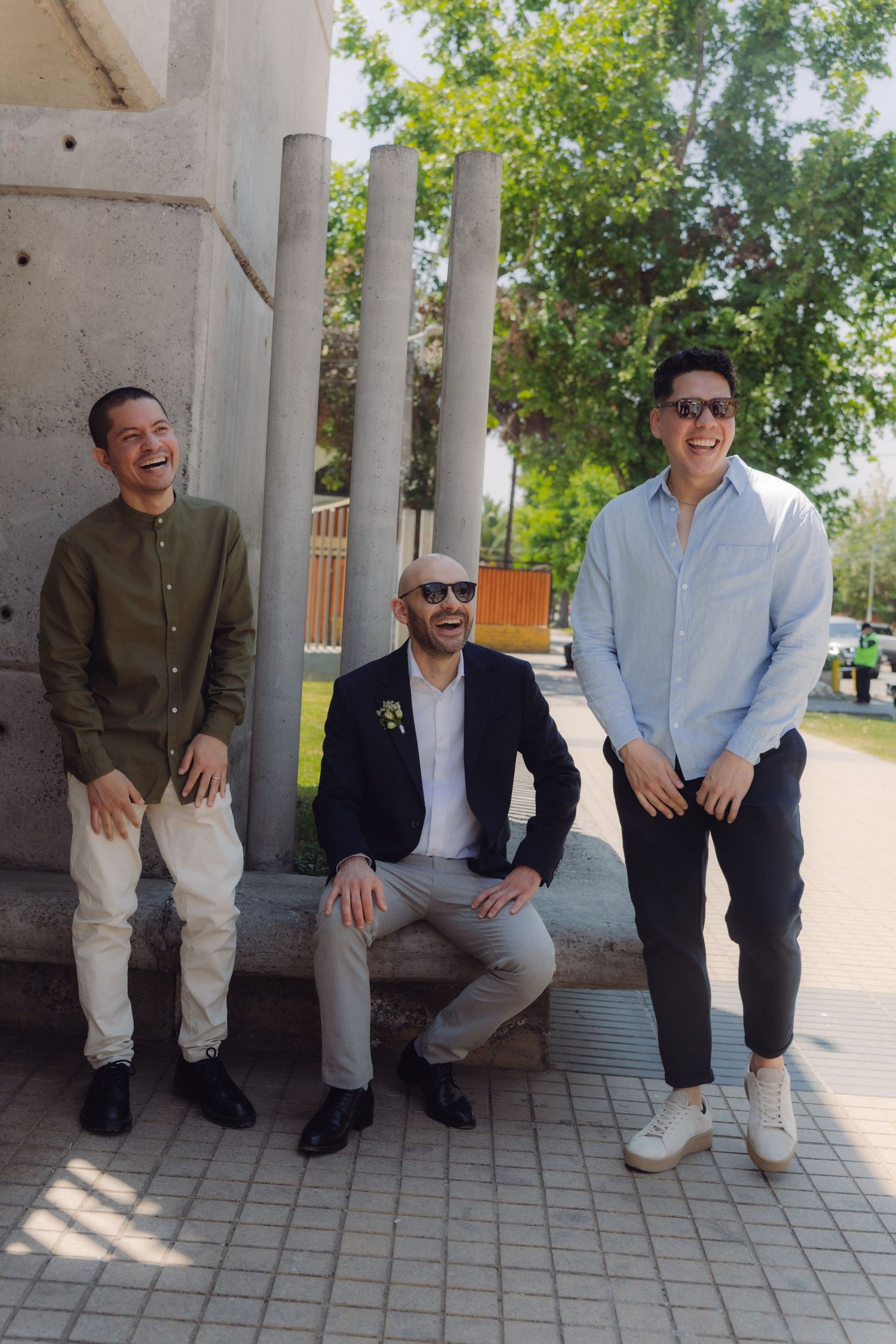 Three smiling men posing outdoors near concrete pillars on a sunny day.
