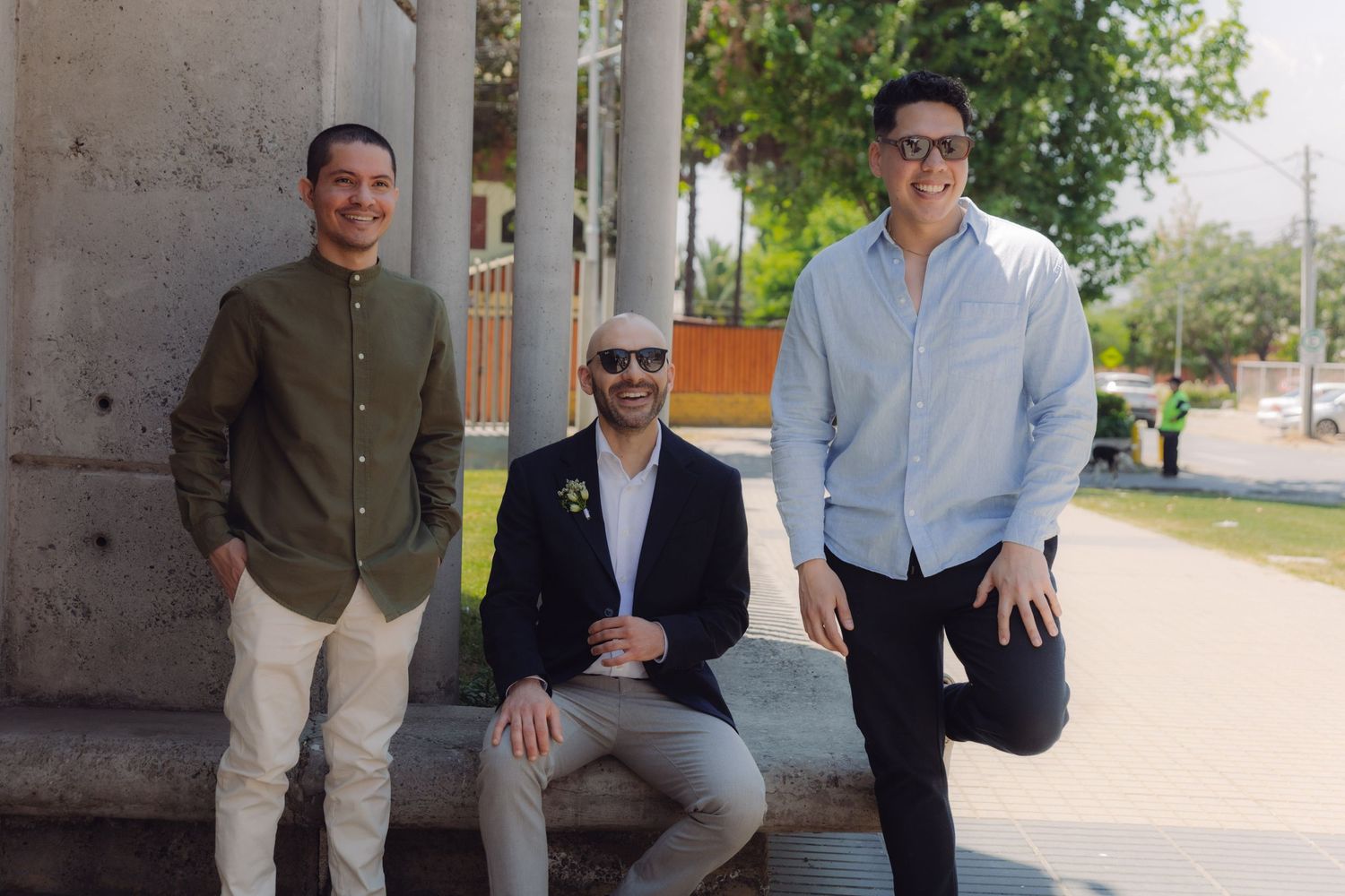 Three men posing casually outdoors near stone columns, smiling on a bright sunny day.
