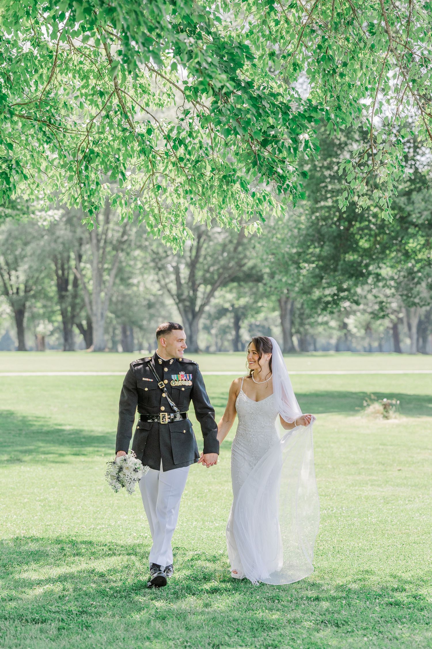 Military groom in dress uniform and bride in white gown walk hand in hand through lush green park on their wedding day.