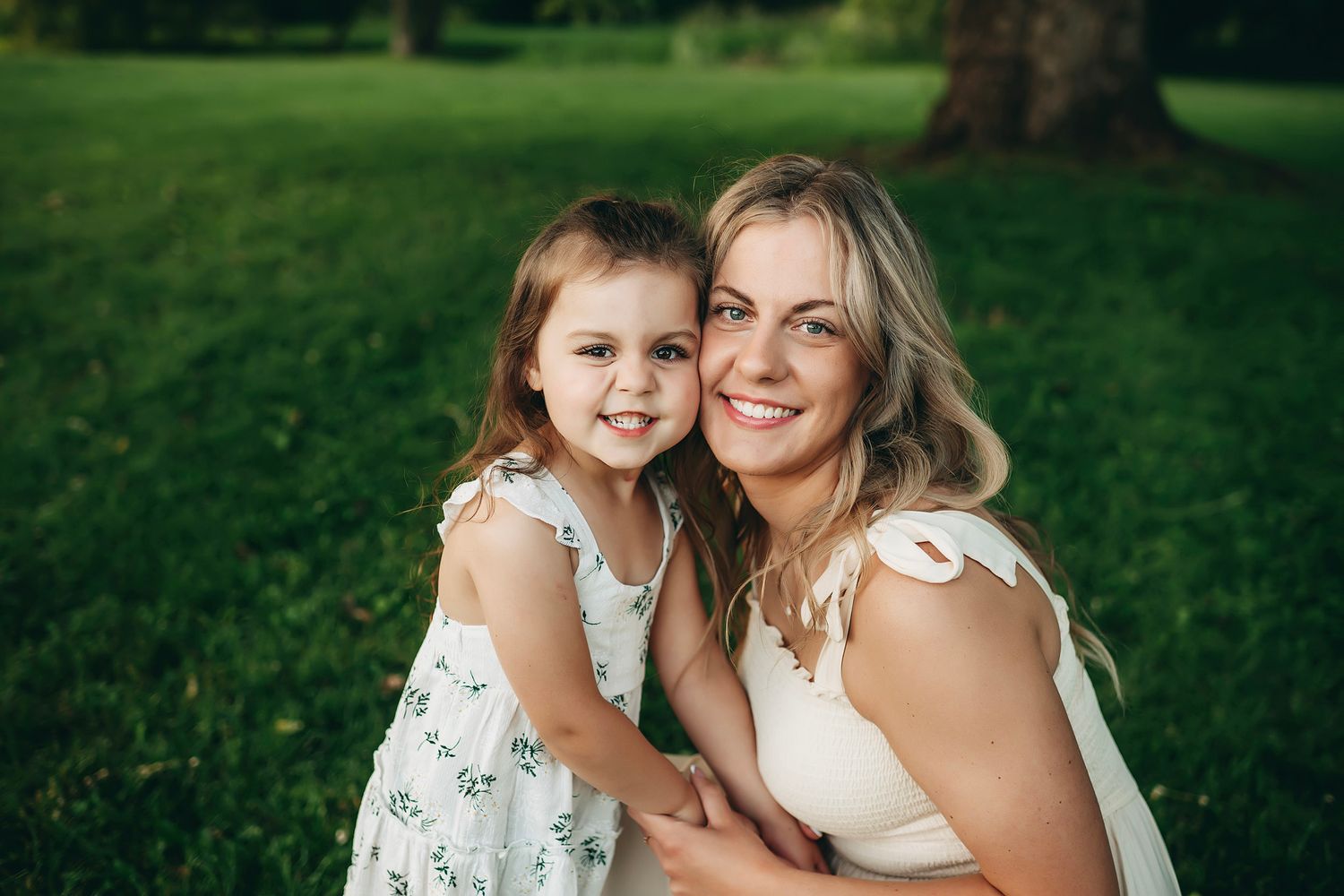 A smiling mother and young daughter in floral dress share a warm embrace on green grass in a sunny park.