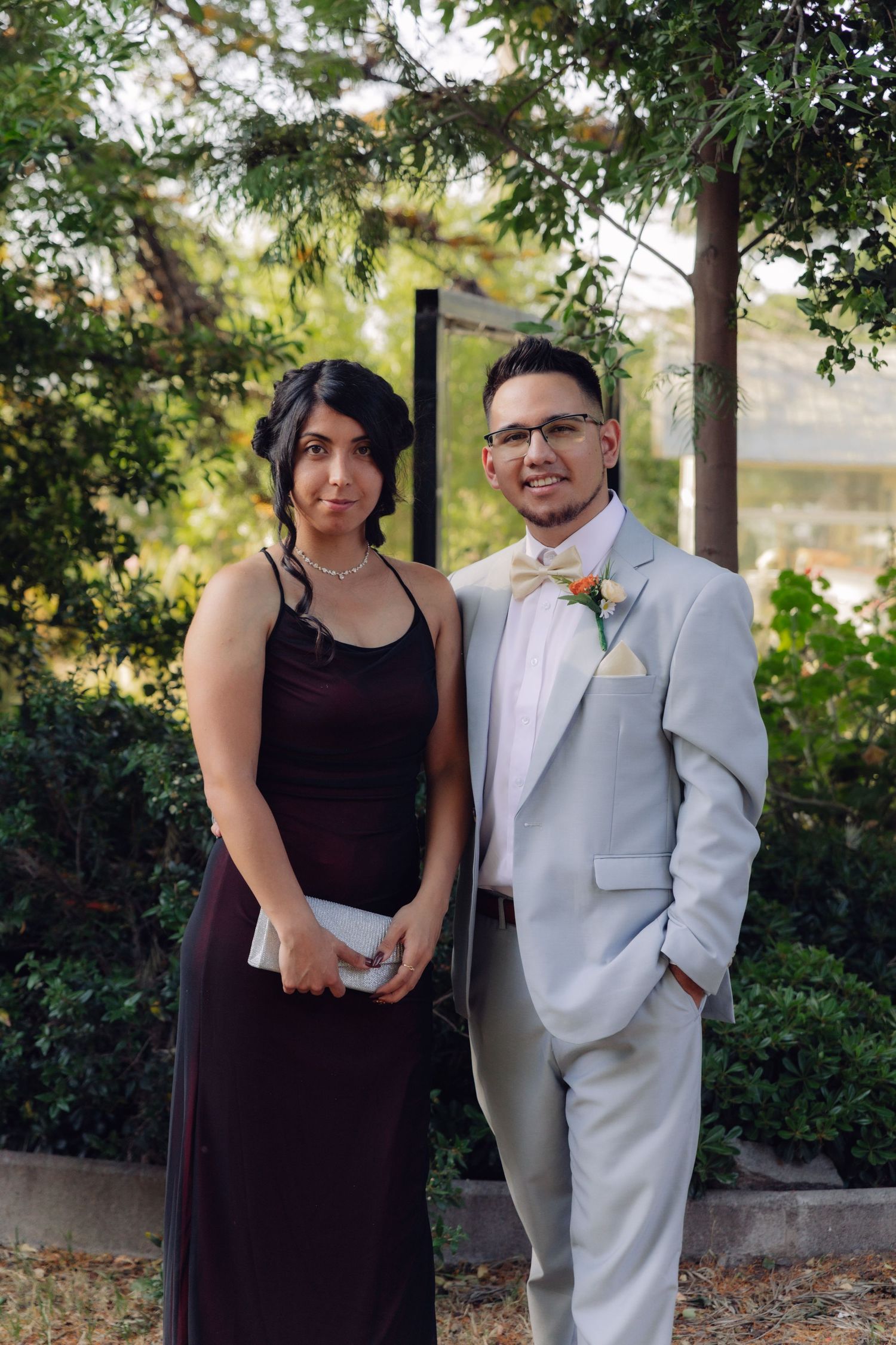 Wedding guest in gray suit with bow tie poses with woman in burgundy gown outdoors at garden venue.