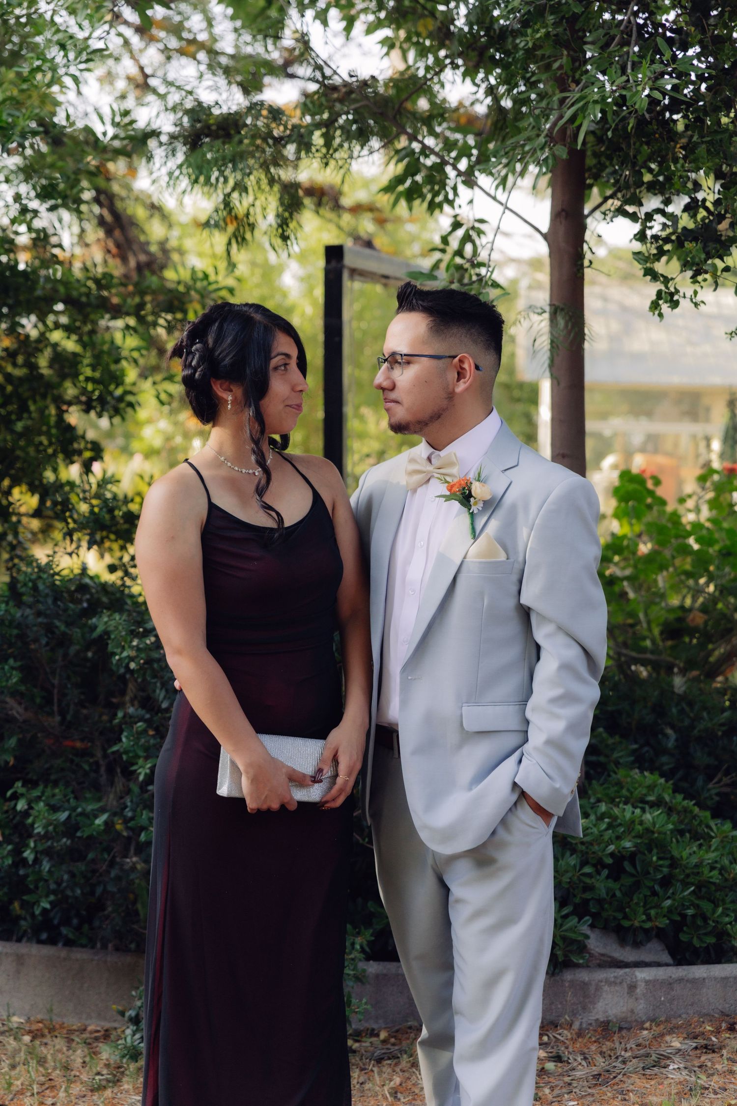 Elegantly dressed couple in gray suit and burgundy gown share a glance at outdoor wedding garden setting.