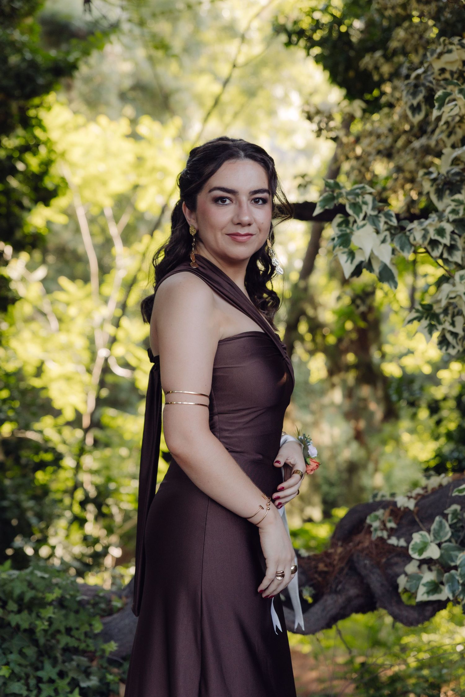 Young woman in brown halter gown poses gracefully among lush green trees at outdoor wedding venue.