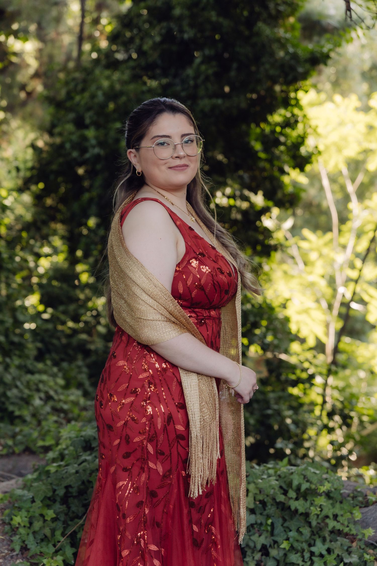 Woman in red floral gown with gold fringe shawl and glasses smiles in sunlit garden wedding setting.