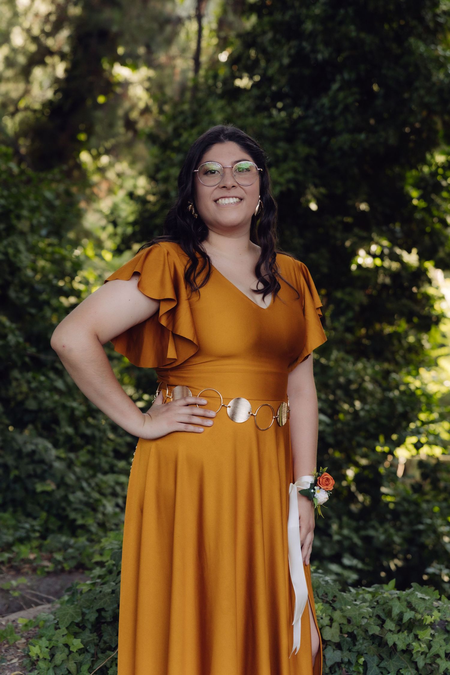 Woman in mustard yellow ruffle dress with gold belt and corsage smiles confidently in garden setting.