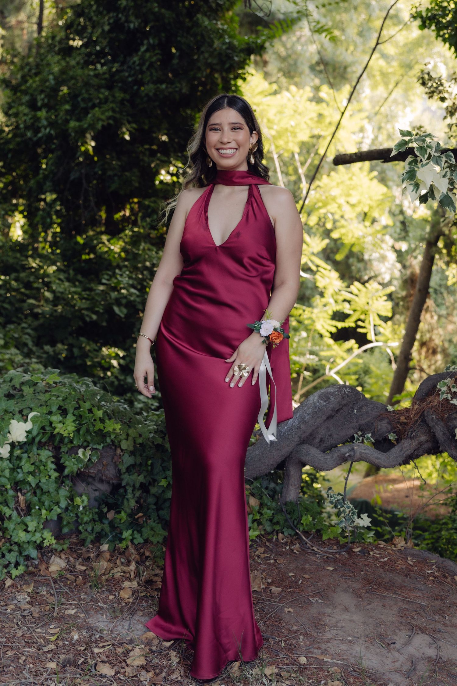 Woman in elegant burgundy satin halter gown posing outdoors in lush green garden setting.