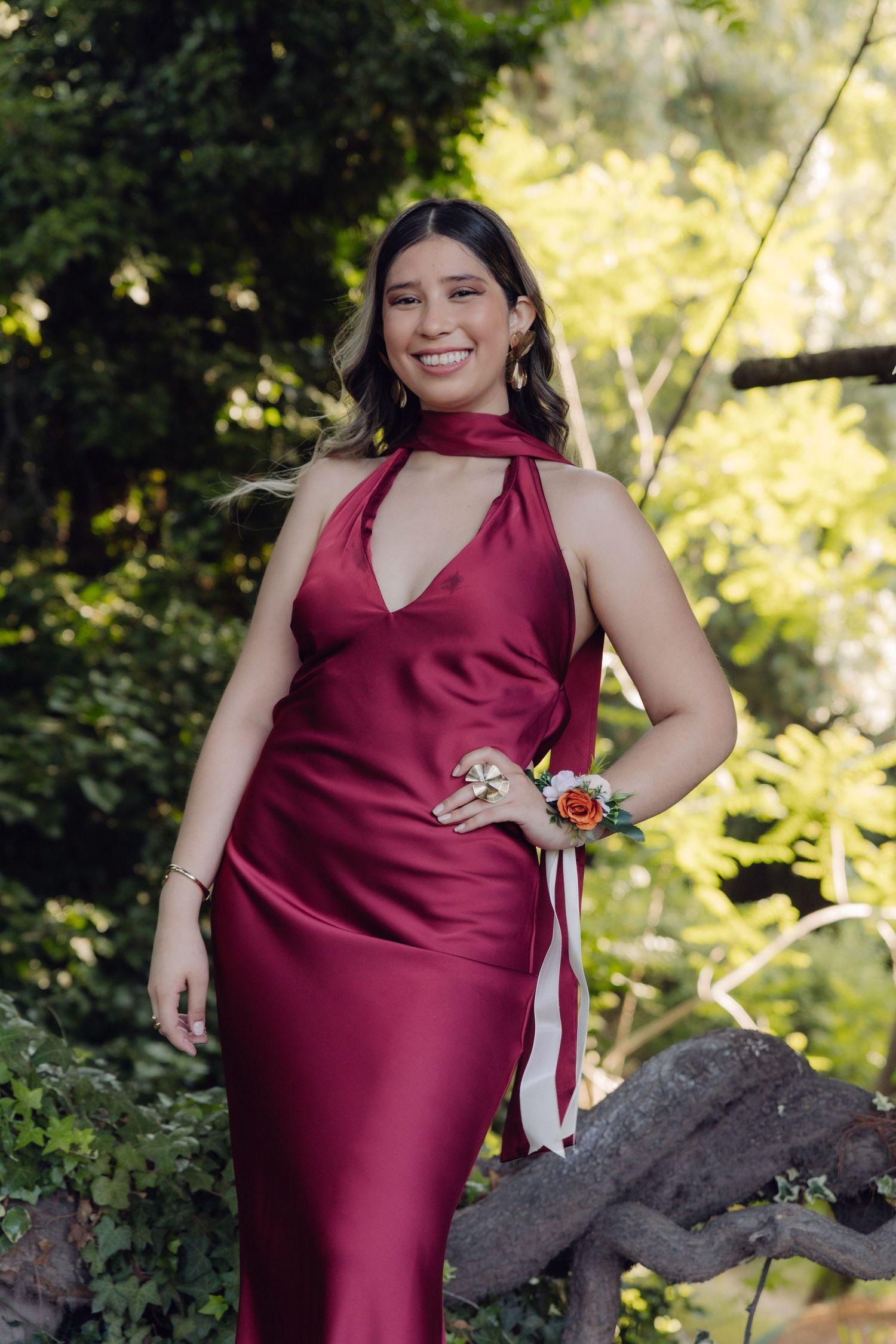 Smiling woman in burgundy satin halter dress with floral wrist corsage in garden.