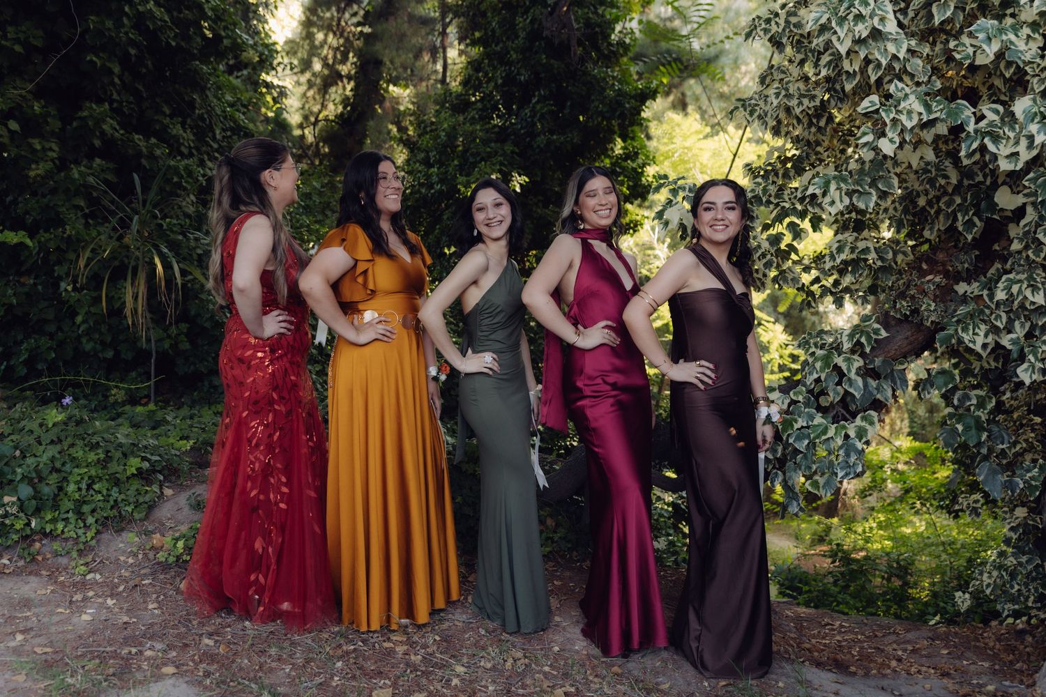 Five women posing in colorful formal gowns in red, gold, burgundy and brown outdoors.