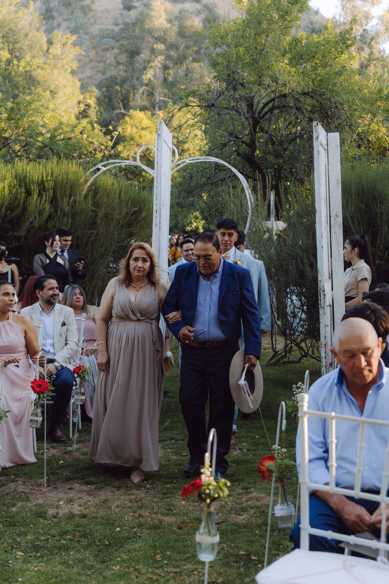 Couple walking down outdoor wedding ceremony aisle with guests seated on white chairs.