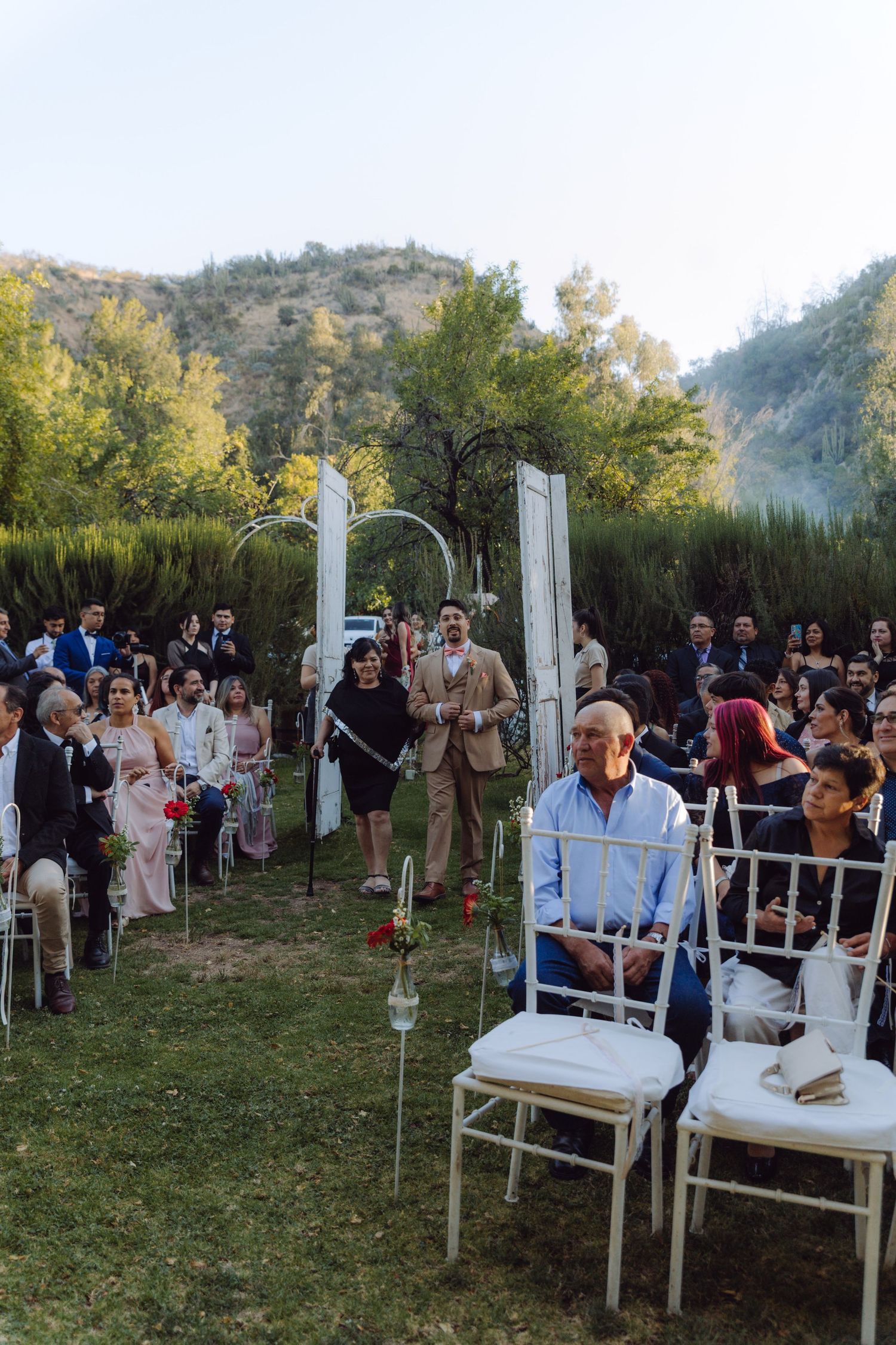 Outdoor wedding ceremony with guests seated on white chairs surrounded by lush green trees.