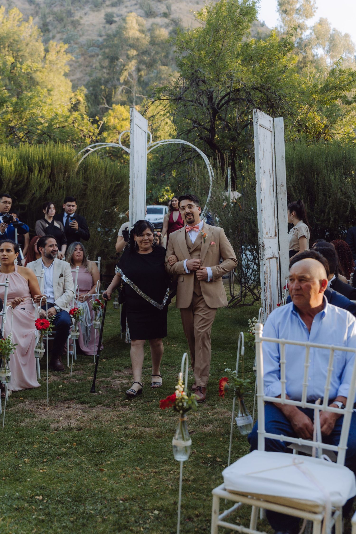 Groom walking down the aisle with his mother at an outdoor garden wedding ceremony.