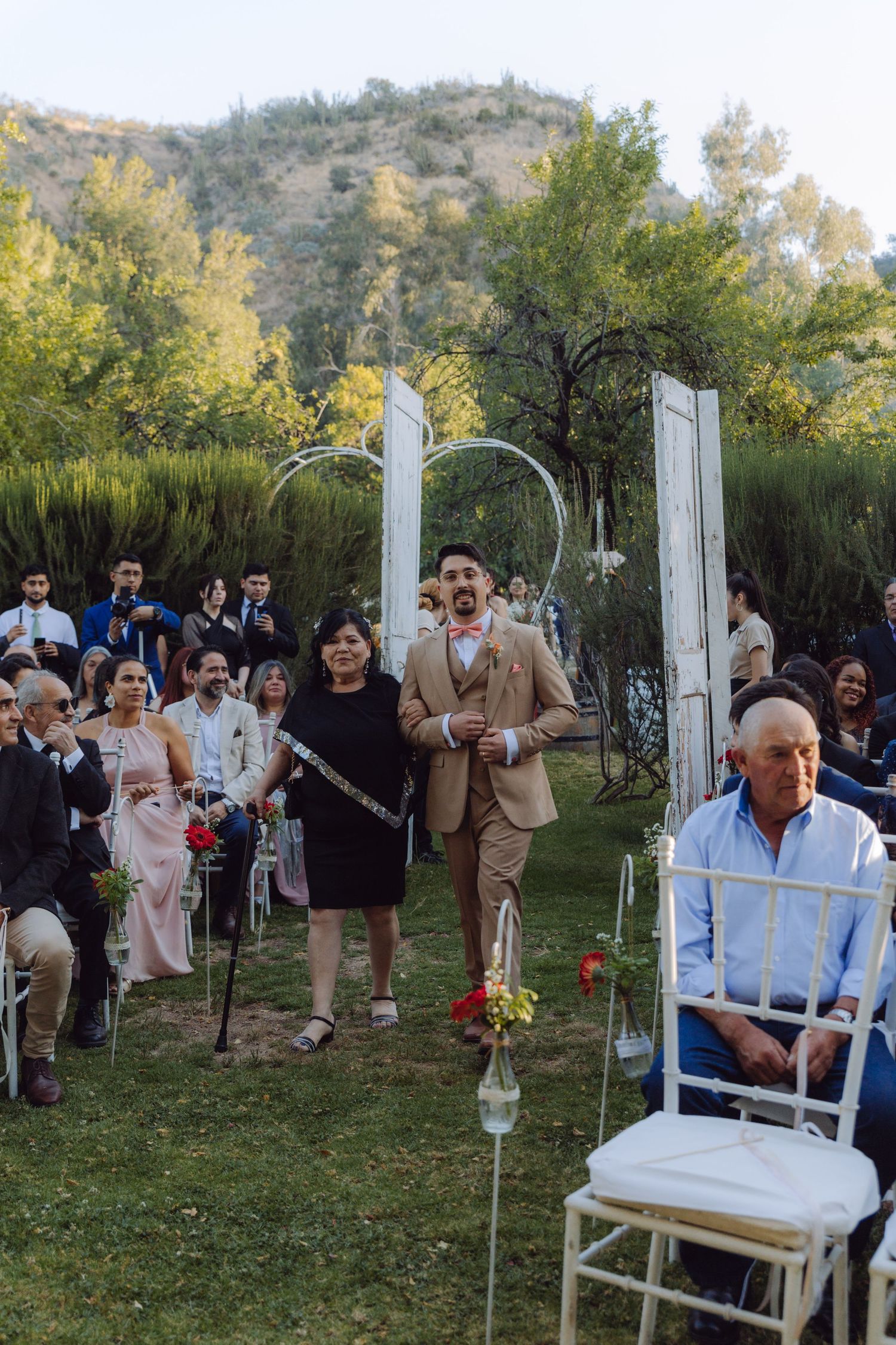 Groom walks down the aisle with family at an outdoor garden wedding ceremony with white arch decor.