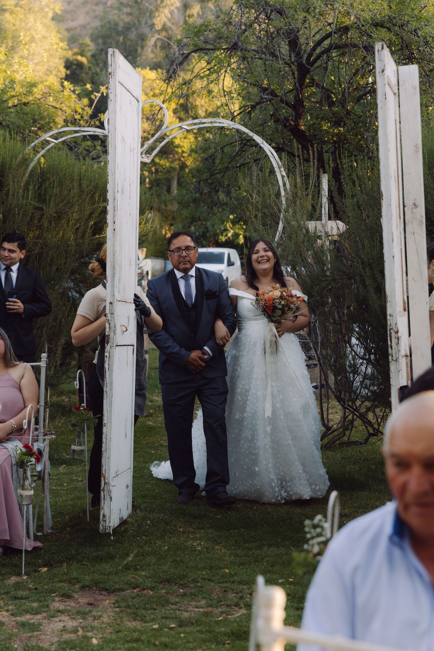 Bride and groom walk down outdoor wedding aisle through white vintage doors surrounded by lush greenery.