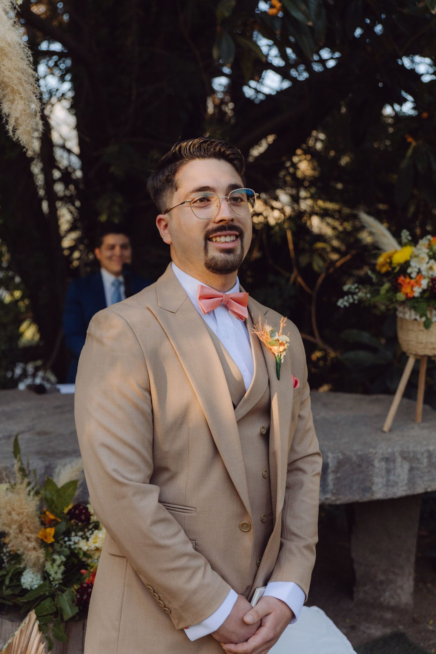 Smiling groom in tan suit with pink bow tie and boutonniere awaits bride at outdoor wedding.