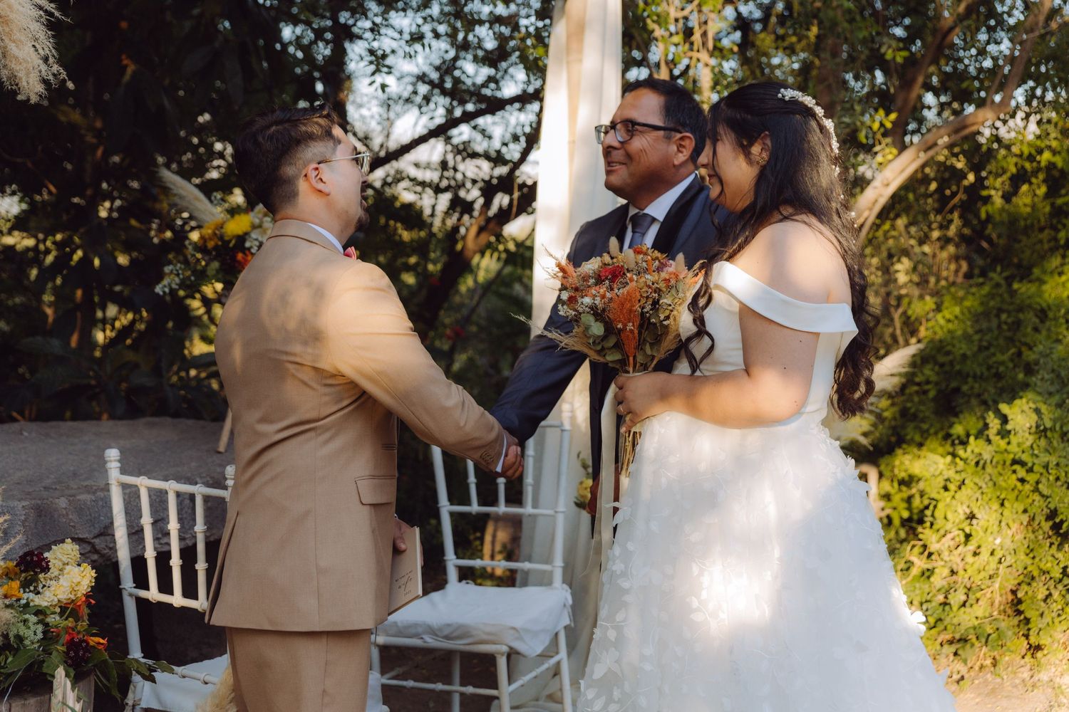 Bride and groom exchange vows at outdoor wedding altar with officiant under floral decorated arch.