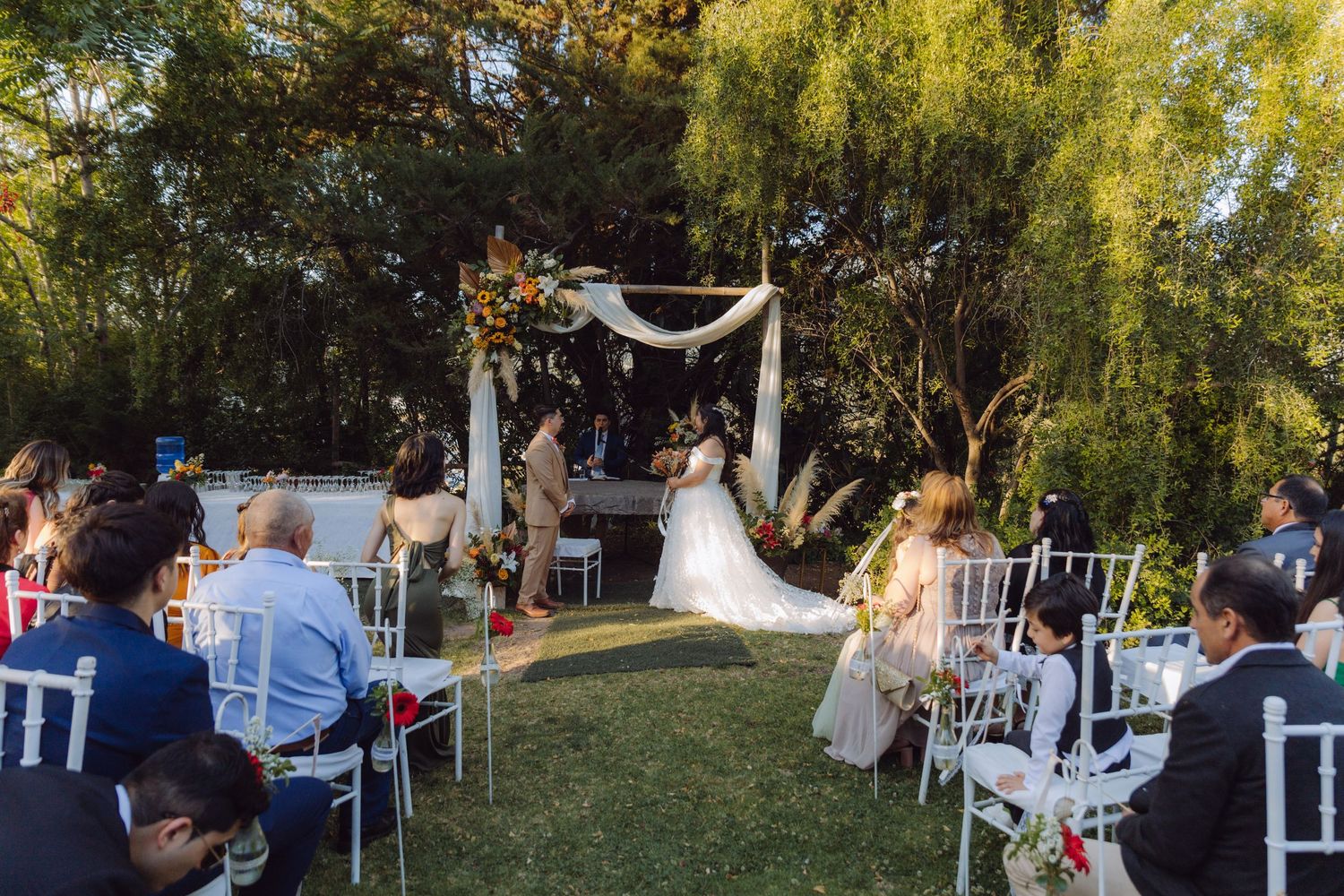 Outdoor garden wedding ceremony with bride and groom at floral arch altar and seated guests.