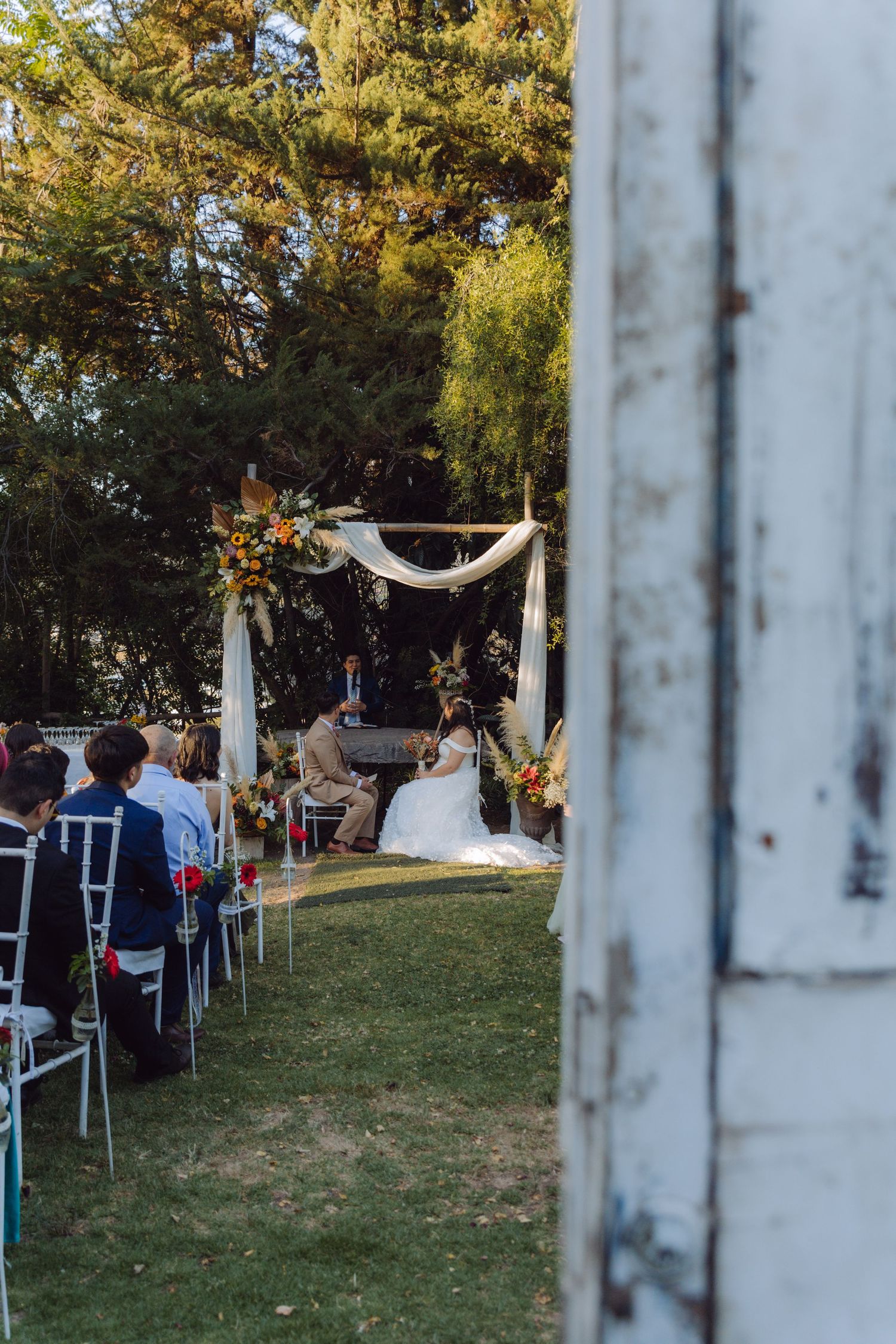 Outdoor wedding ceremony viewed through vintage white door frame with couple at decorated altar.