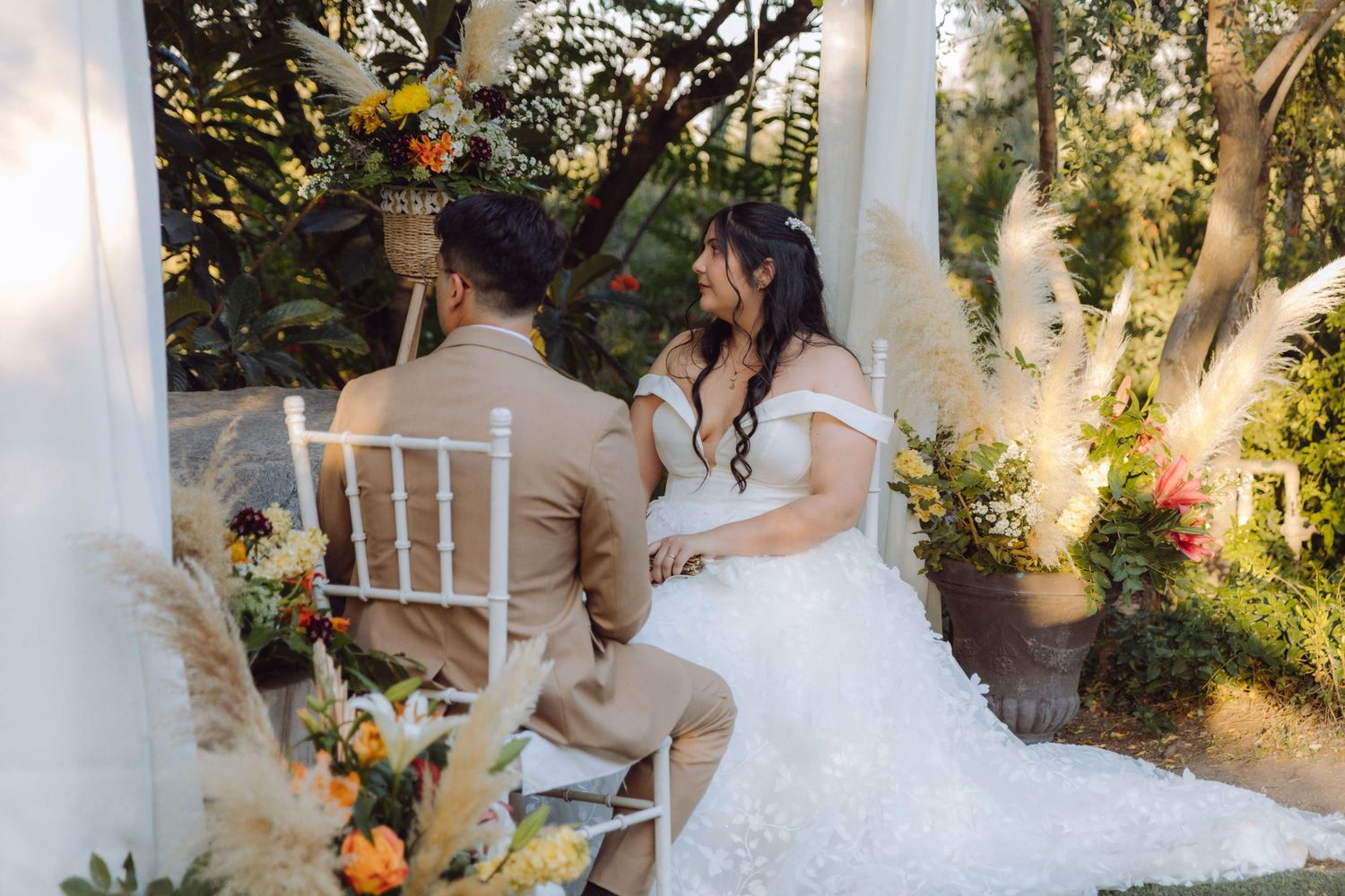 Bride and groom sit together at outdoor wedding altar surrounded by lush greenery and floral arrangements.