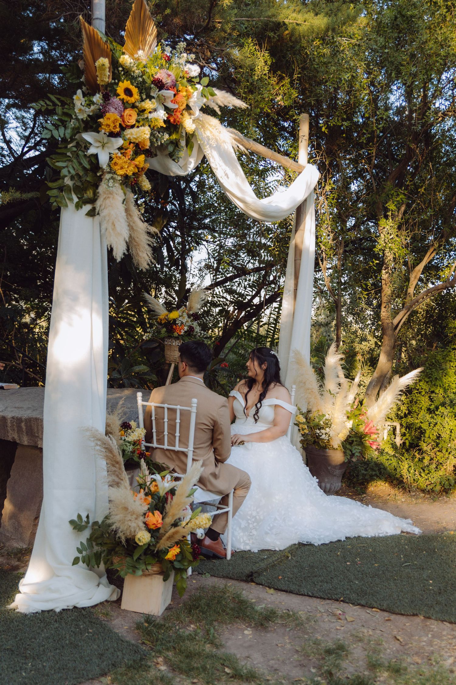 Bride and groom seated under a floral arch adorned with sunflowers and white draping at outdoor wedding.