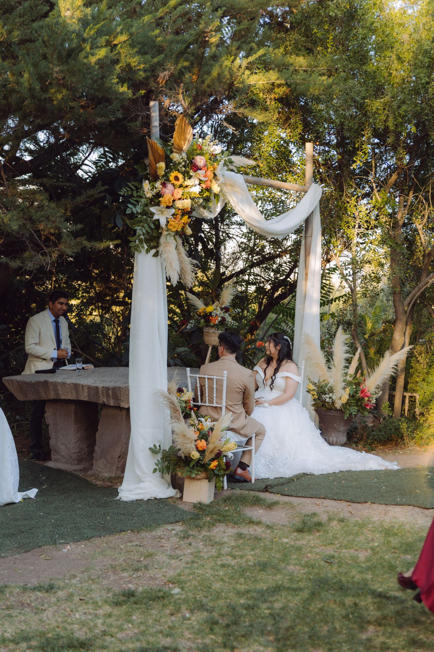 Wedding couple seated under decorated floral arch while officiant reads ceremony at outdoor garden wedding.