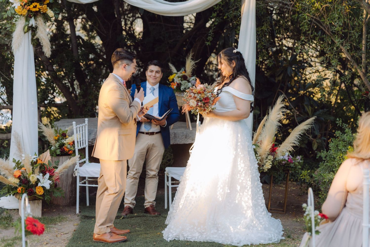 Groom reads vows to bride while officiant stands behind them at a boho outdoor garden wedding ceremony.