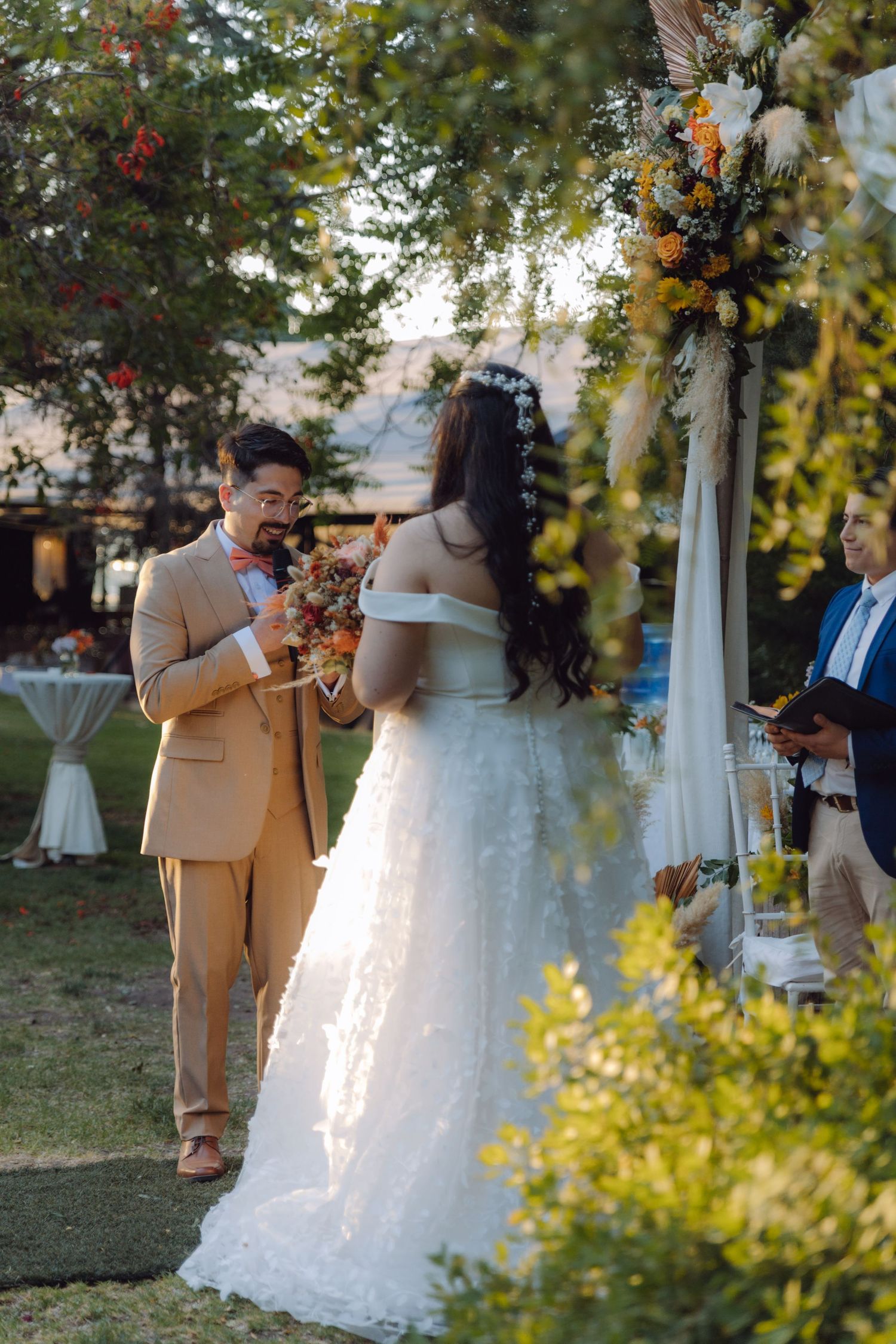 Groom in tan suit reads vows to bride in white off-shoulder gown at outdoor wedding ceremony.