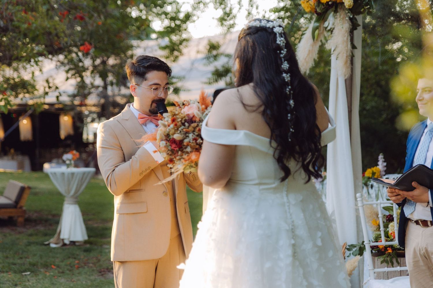 Groom holds colorful bridal bouquet facing bride at a sunlit outdoor garden wedding ceremony.