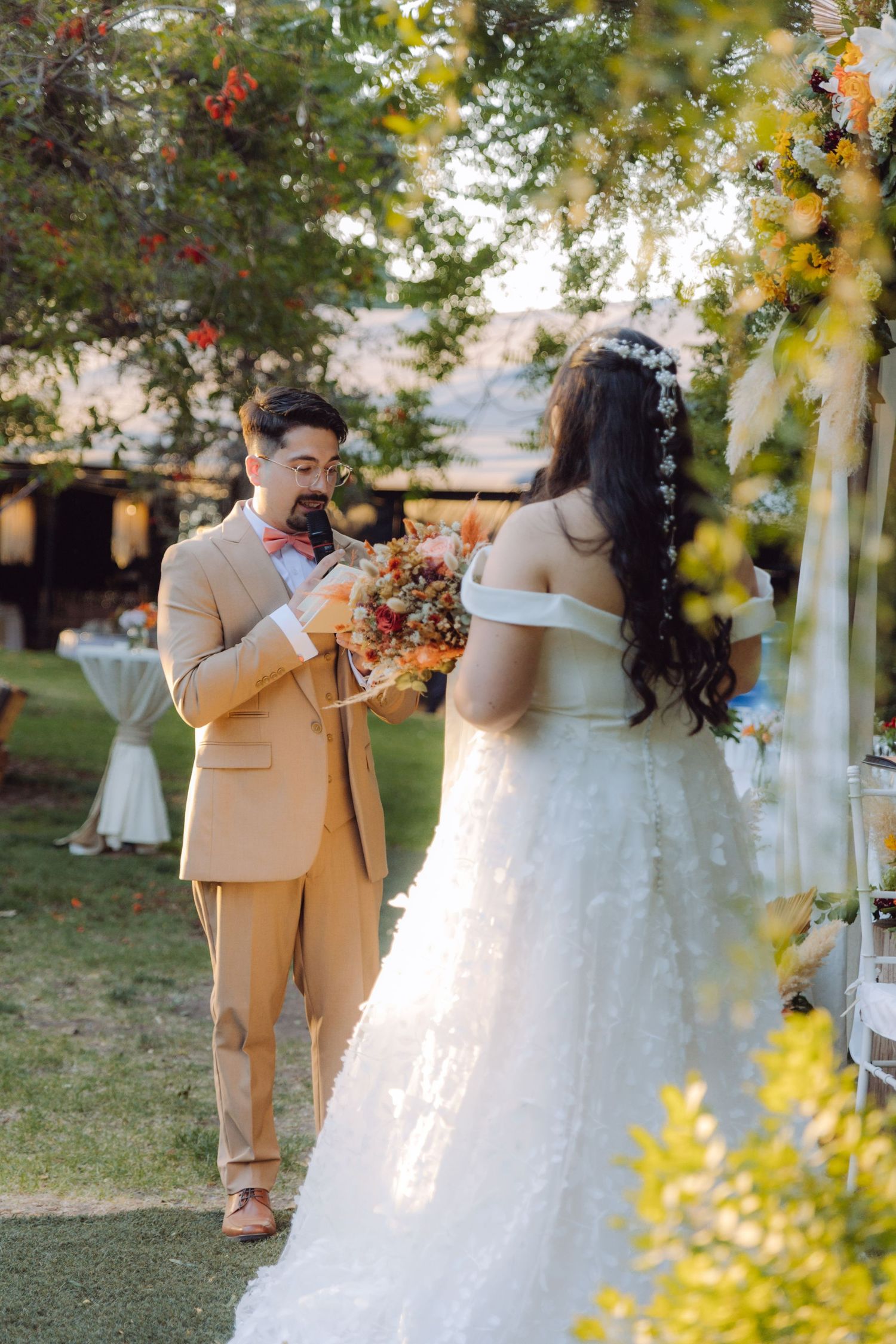 Groom kisses bridal bouquet of autumn flowers while facing bride at outdoor garden wedding ceremony.