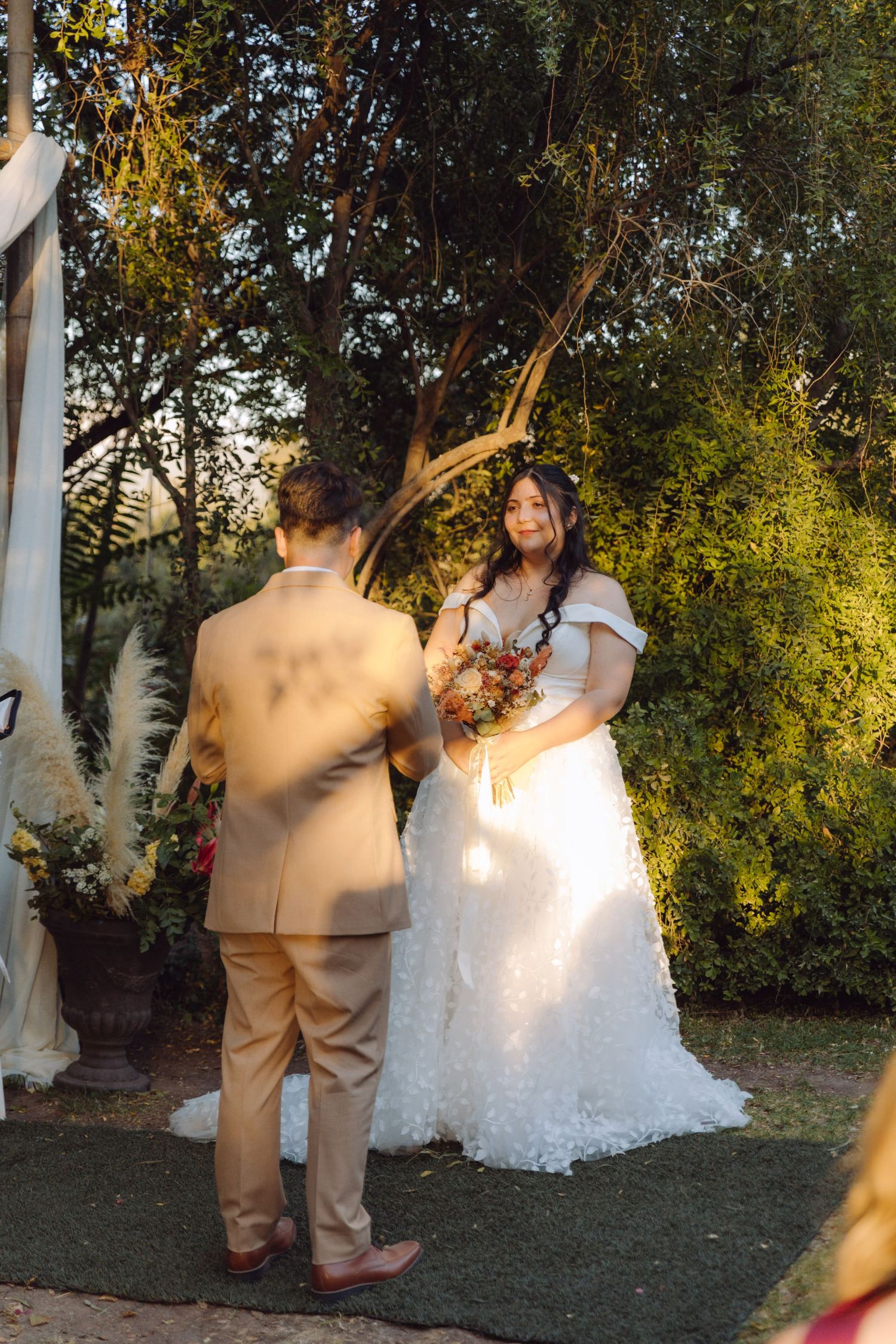 Bride in white lace gown holds bouquet facing groom at golden hour outdoor wedding ceremony.