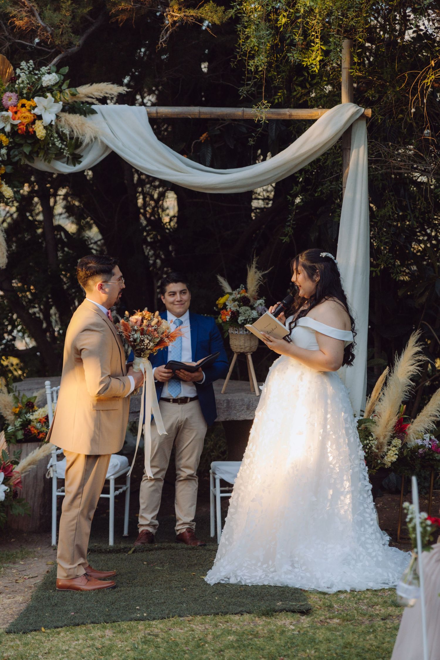 Bride reads vows to groom at outdoor wedding ceremony under wooden arch with white draping and flowers.