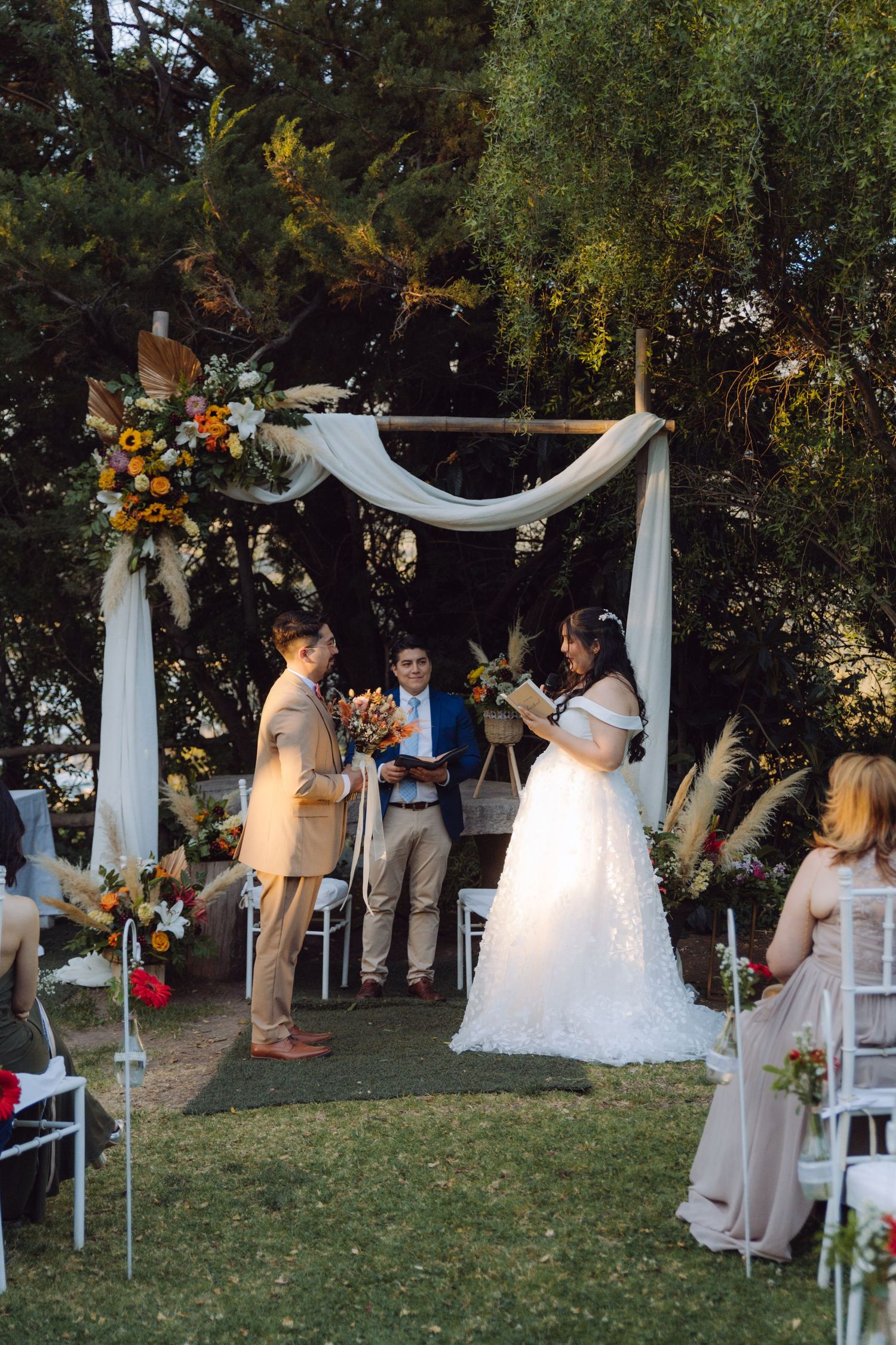 Bride reads vows at outdoor wedding ceremony under rustic wooden arch adorned with colorful flowers.