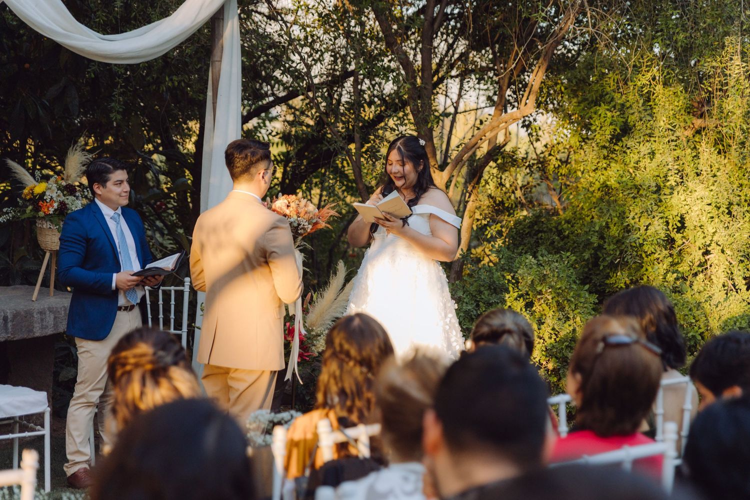 Bride placing wedding ring on groom's finger at outdoor ceremony, officiant in blue suit, guests seated.