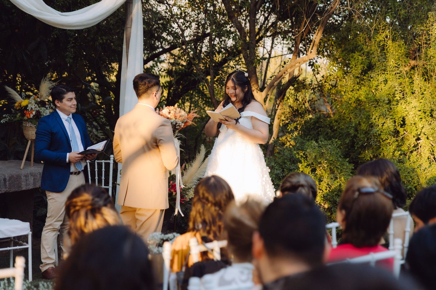 Bride and groom exchange rings at outdoor wedding ceremony as officiant and guests look on.