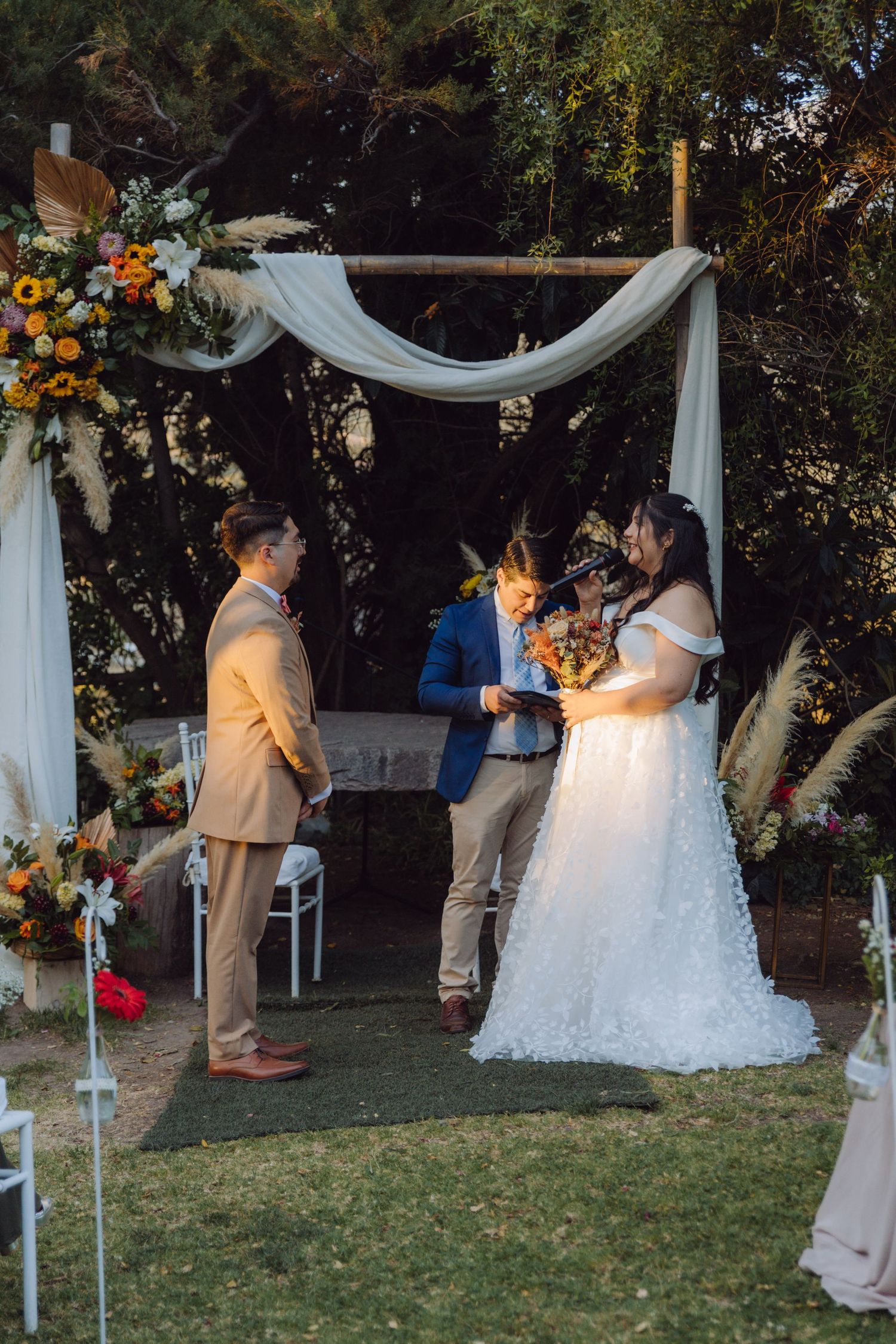 Bride and groom exchange vows under a floral arch with white draping at an outdoor wedding ceremony.