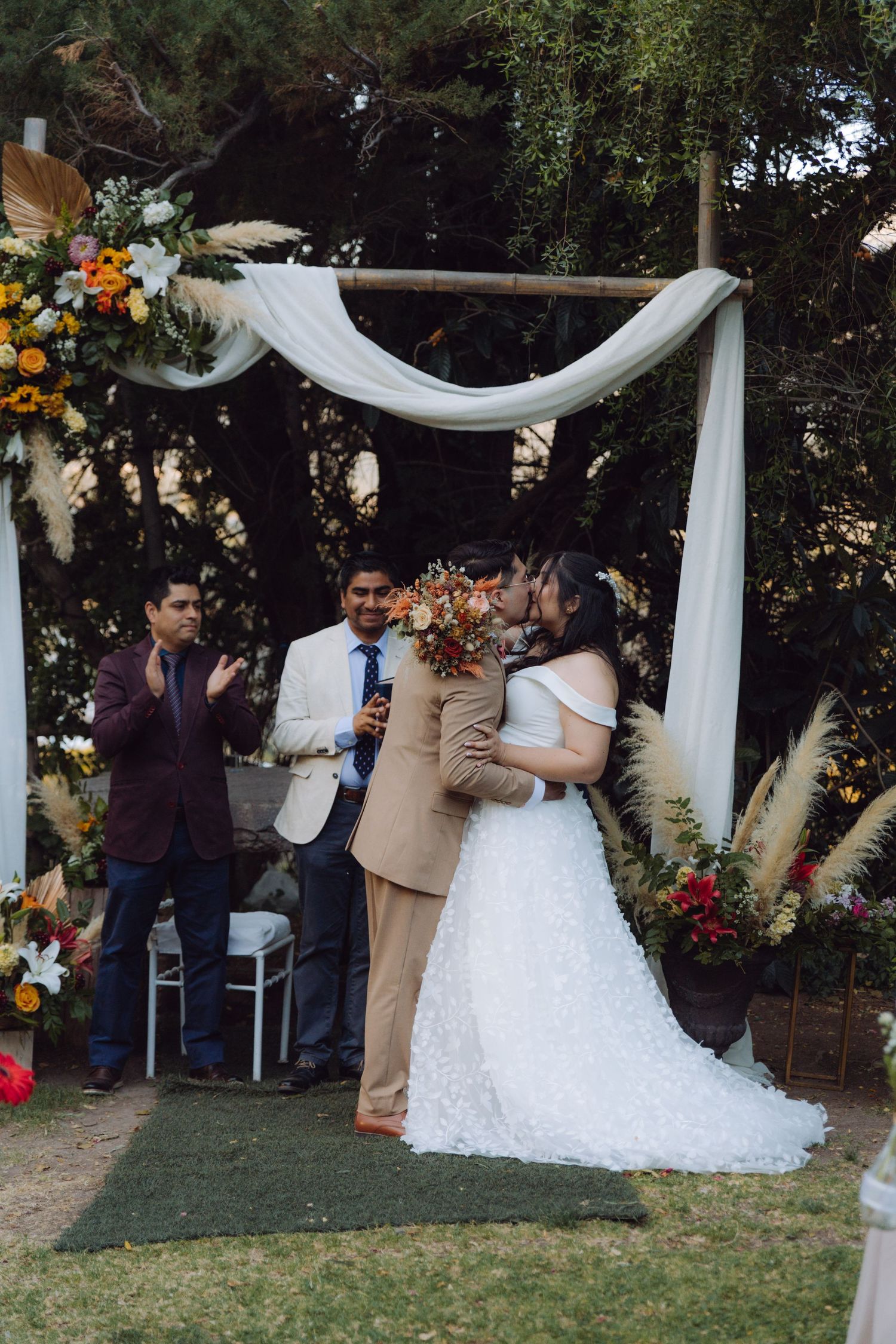 Bride and groom share first kiss under a decorated arch as guests applaud at outdoor wedding.