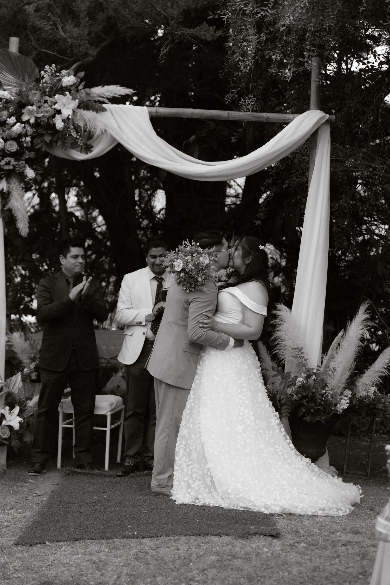 Black and white photo of bride and groom sharing first kiss at outdoor wedding ceremony under floral arch.