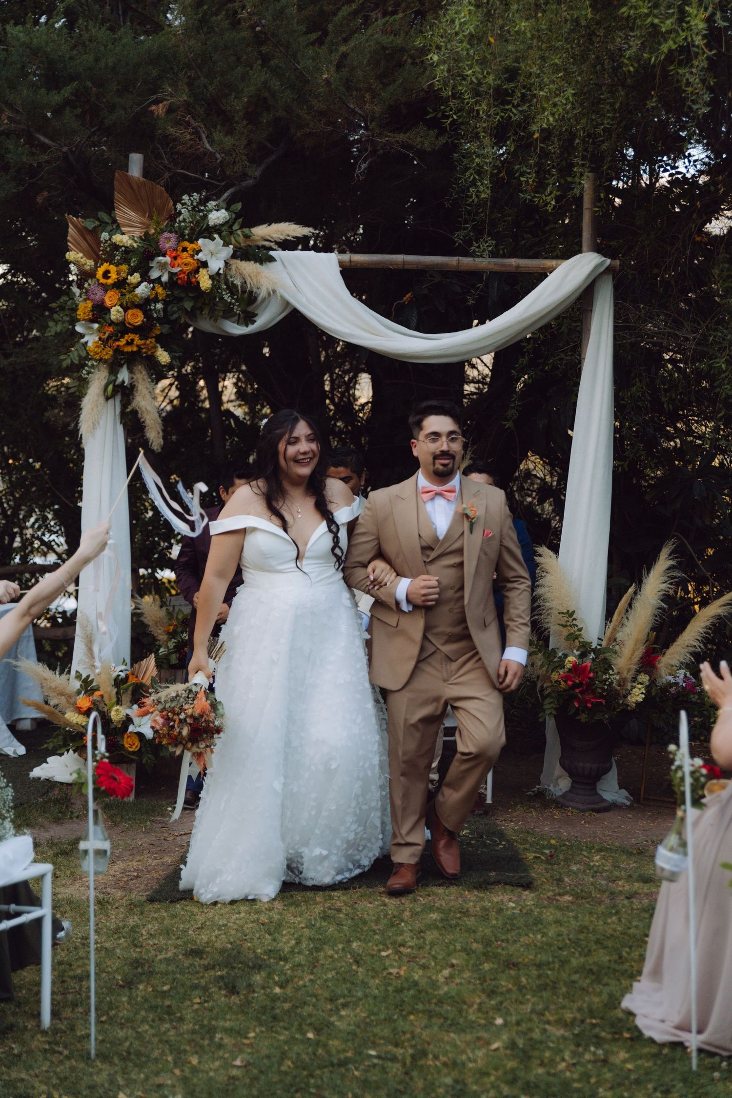 Smiling newlyweds walk down aisle after outdoor wedding ceremony under decorated wooden arch with flowers.